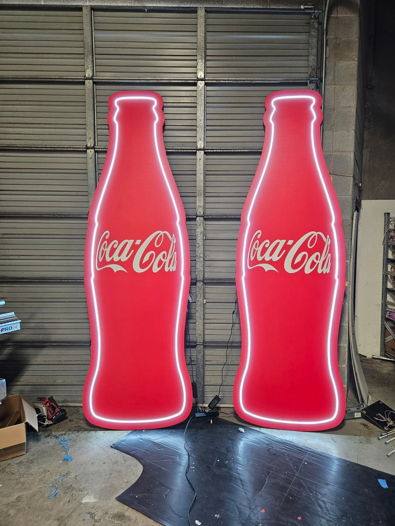 Two oversized Neon Coca-Cola bottle signs with lit outline, positioned indoors in front of a metal rolling garage door. The signs are red with white Coca-Cola logos, and there are various tools and boxes on the garage floor.
