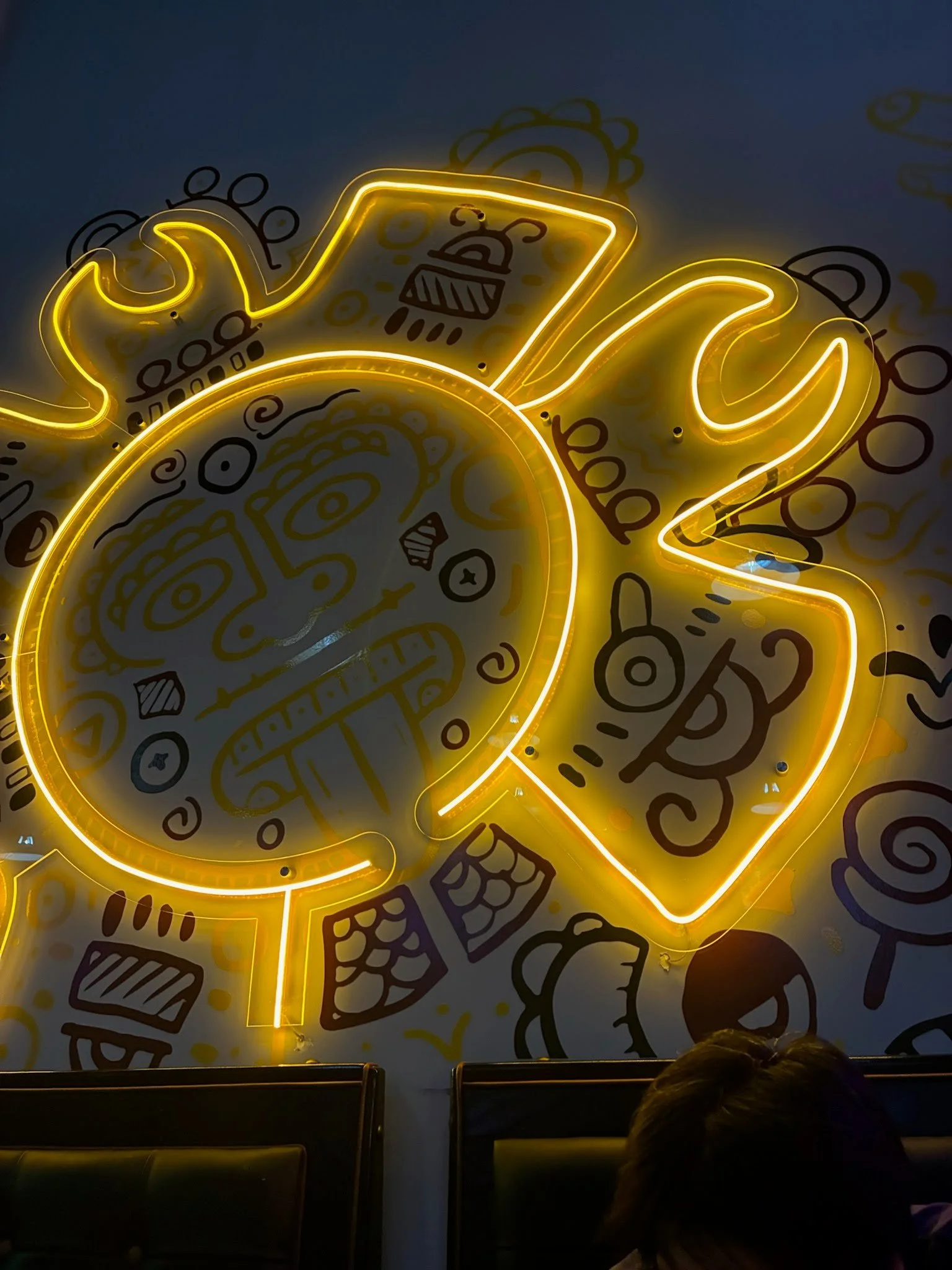 Neon sign of a taco with yellow outline, mounted on a dark wall with doodle-style illustrations of food and kitchen items. Part of a seated audience visible at the bottom.