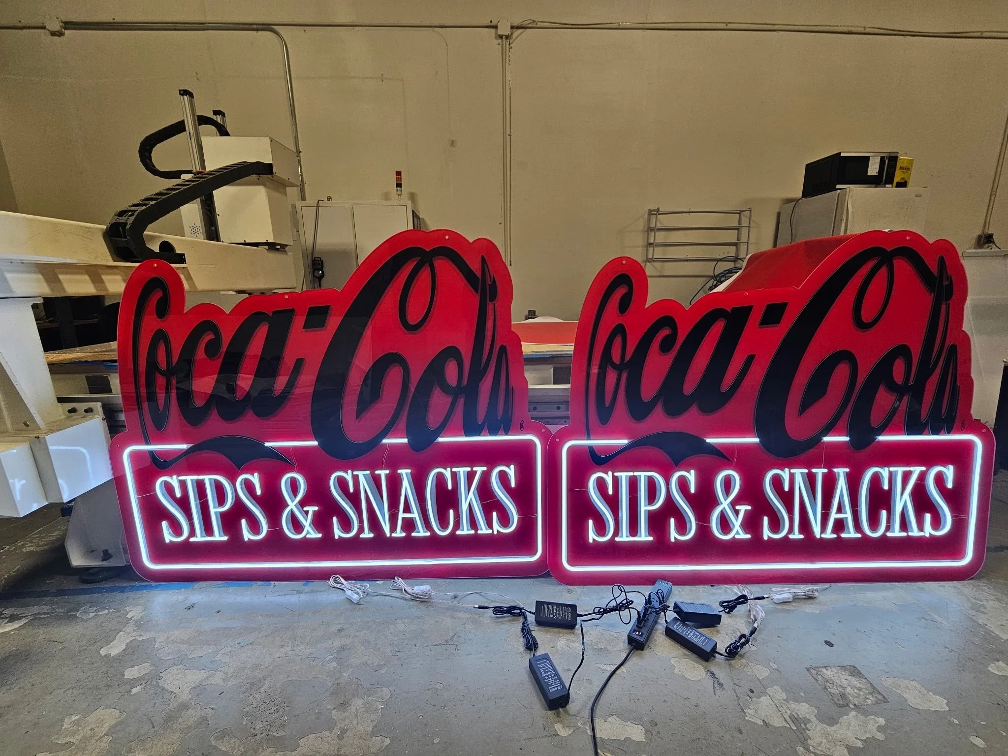 LED signs illuminated with text reading 'SIPS & SNACKS' in pink and white colors, placed on a concrete floor with electronic equipment and cords nearby; background includes a plain wall with some industrial items.