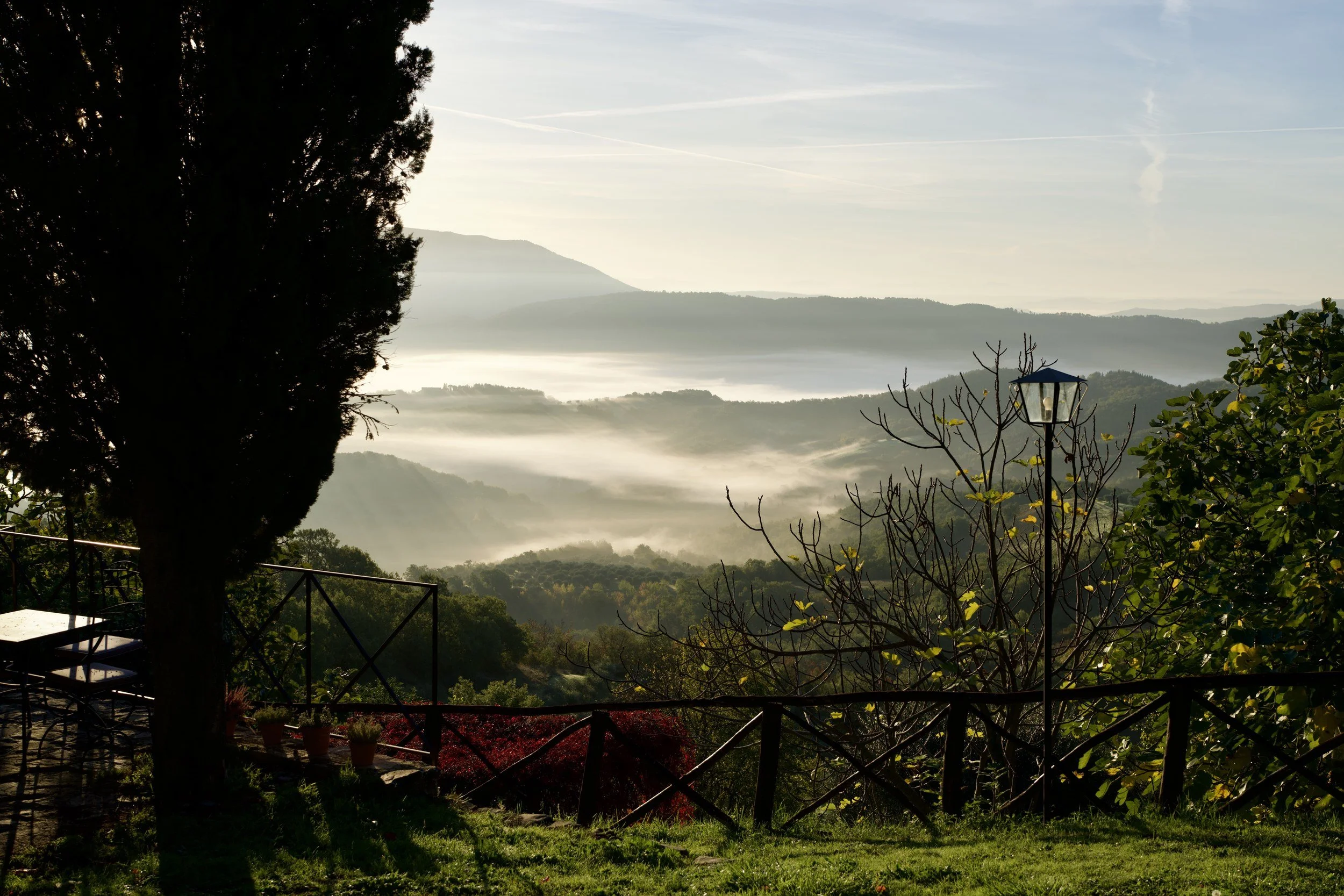 Morning fog filling the valleys below wooded hills, seen past a cypress tree and a red fence in the Italian countryside.