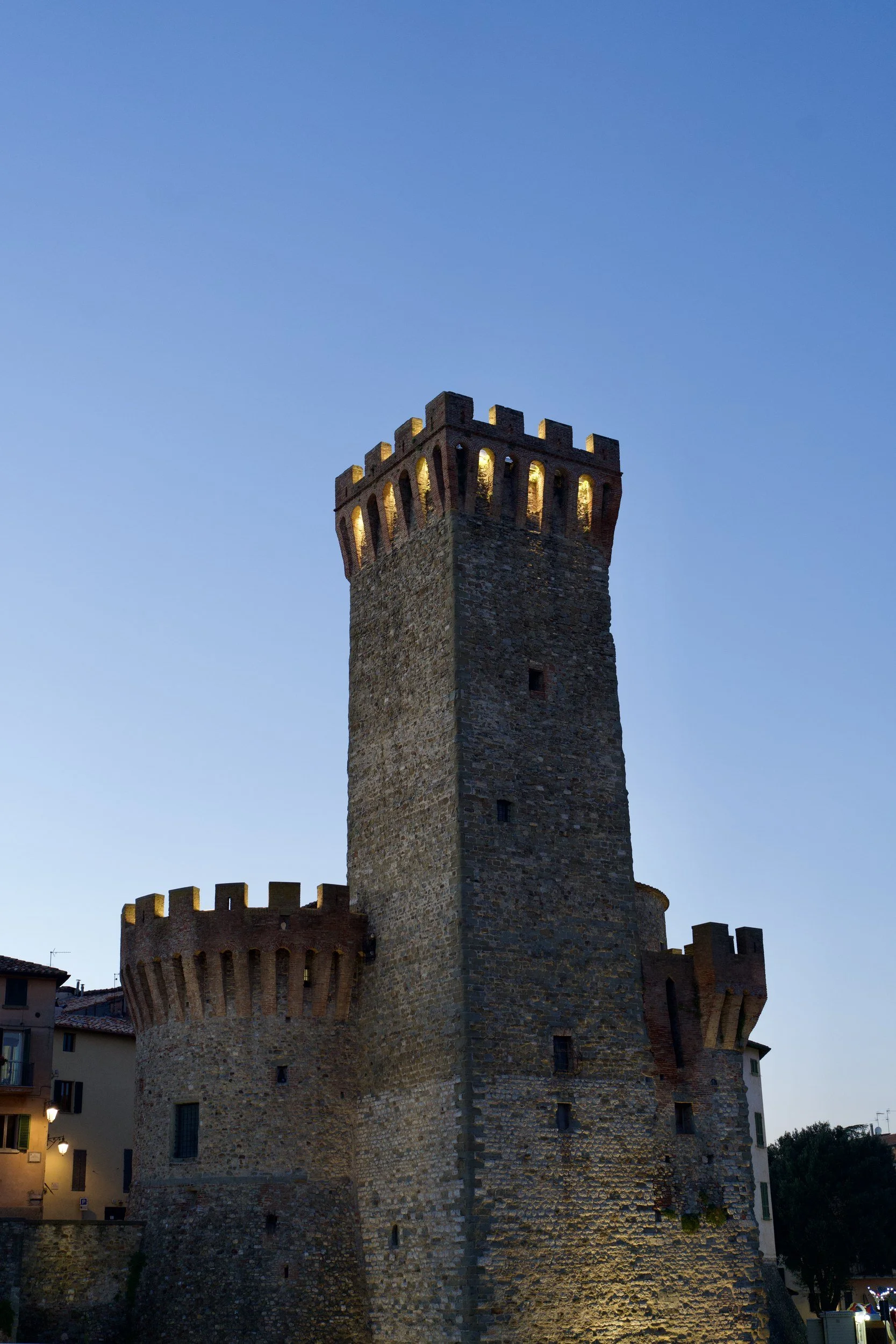 A crenellated medieval stone tower and castle wall against a clear blue sky in Italy.