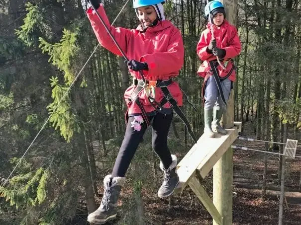 Two children in red jackets and helmets walking on a wooden balance beam at a high ropes course in a forest.