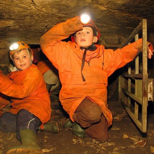 Two children in orange caving helmets and jackets exploring a cave. One is holding a flashlight and the other is near a metal gate, with dirt and leaves on the ground.