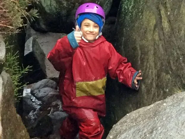 Child wearing red rain gear and a purple helmet, giving a thumbs up while exploring between rocks.