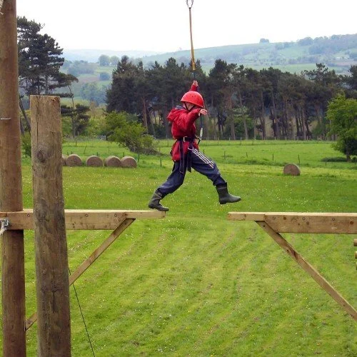 Child in safety gear walking on a wooden obstacle course in an outdoor park.