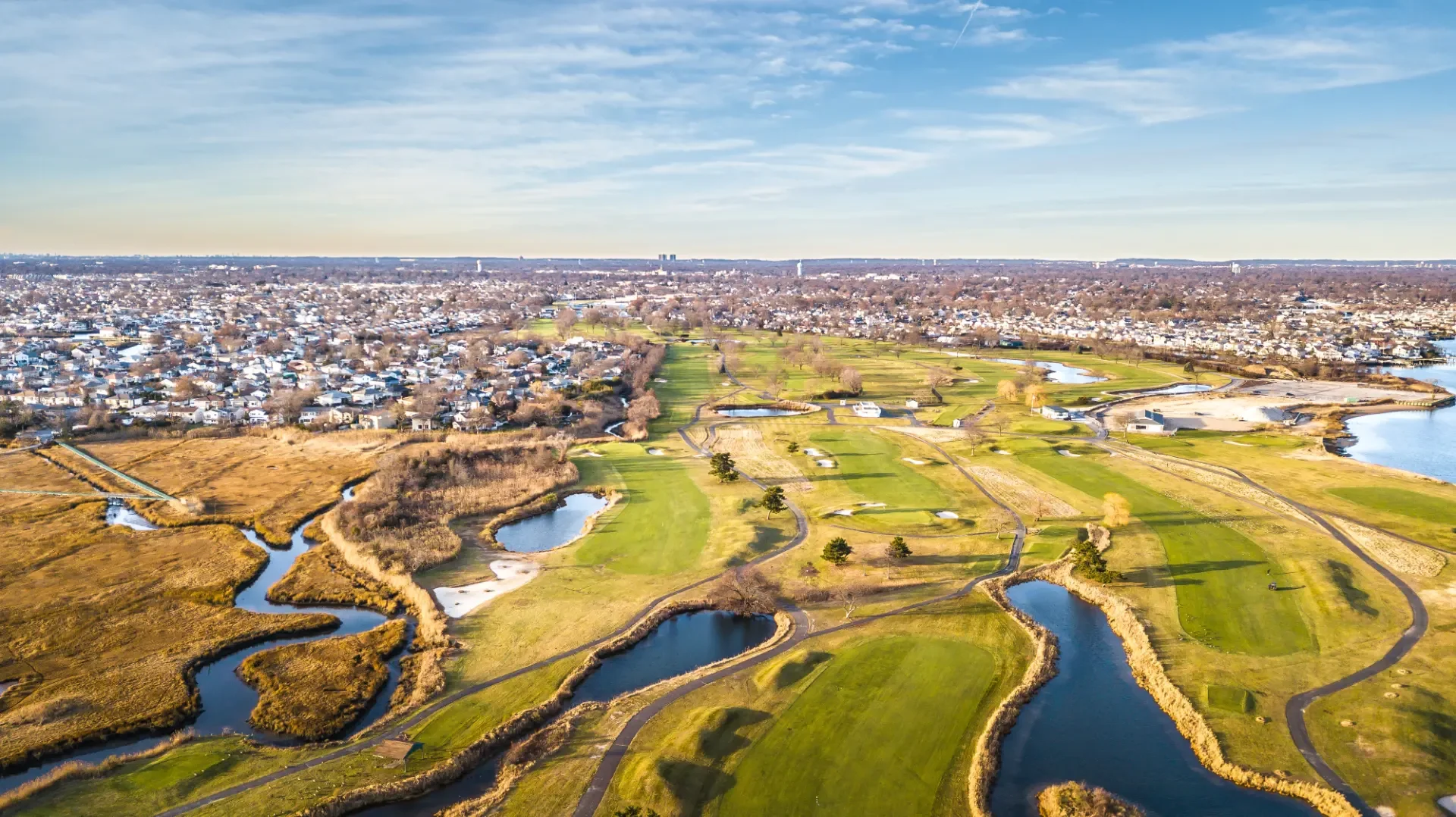 Aerial view of a golf course with water hazards and a nearby residential area under a partly cloudy sky.