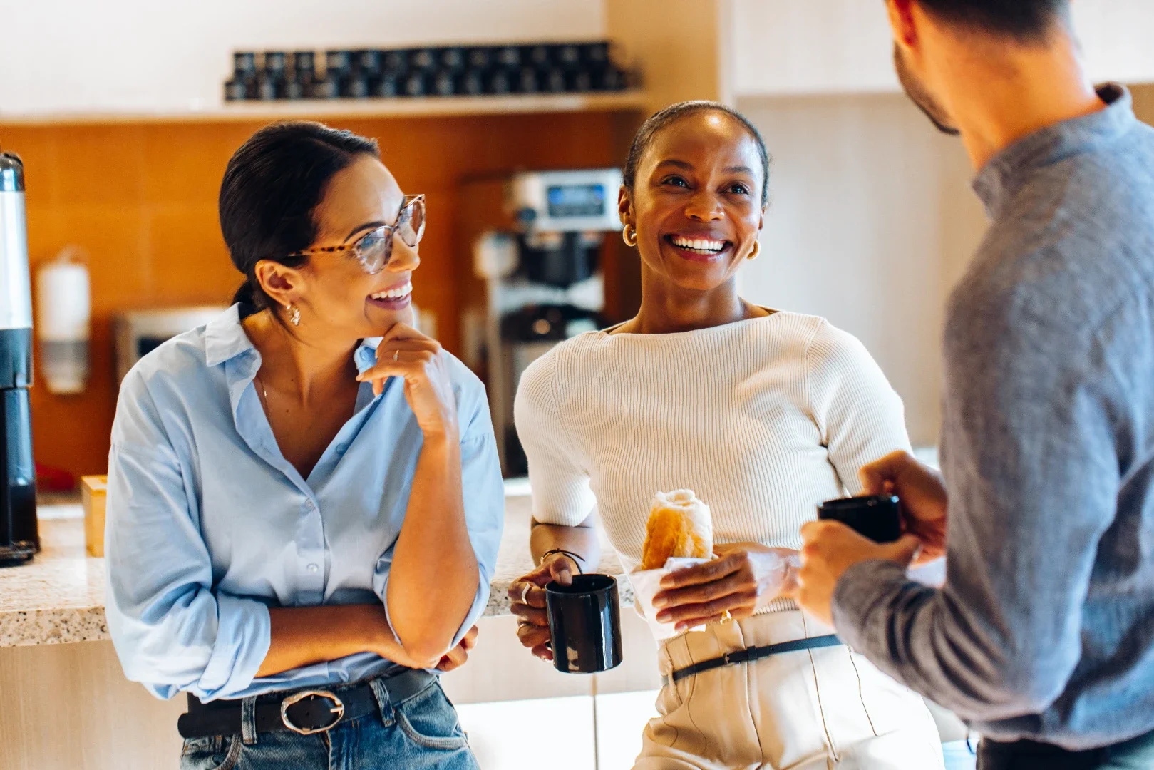 Three women in a kitchen having a conversation, one holding a coffee mug and a pastry, smiling and engaging with a man holding a phone.