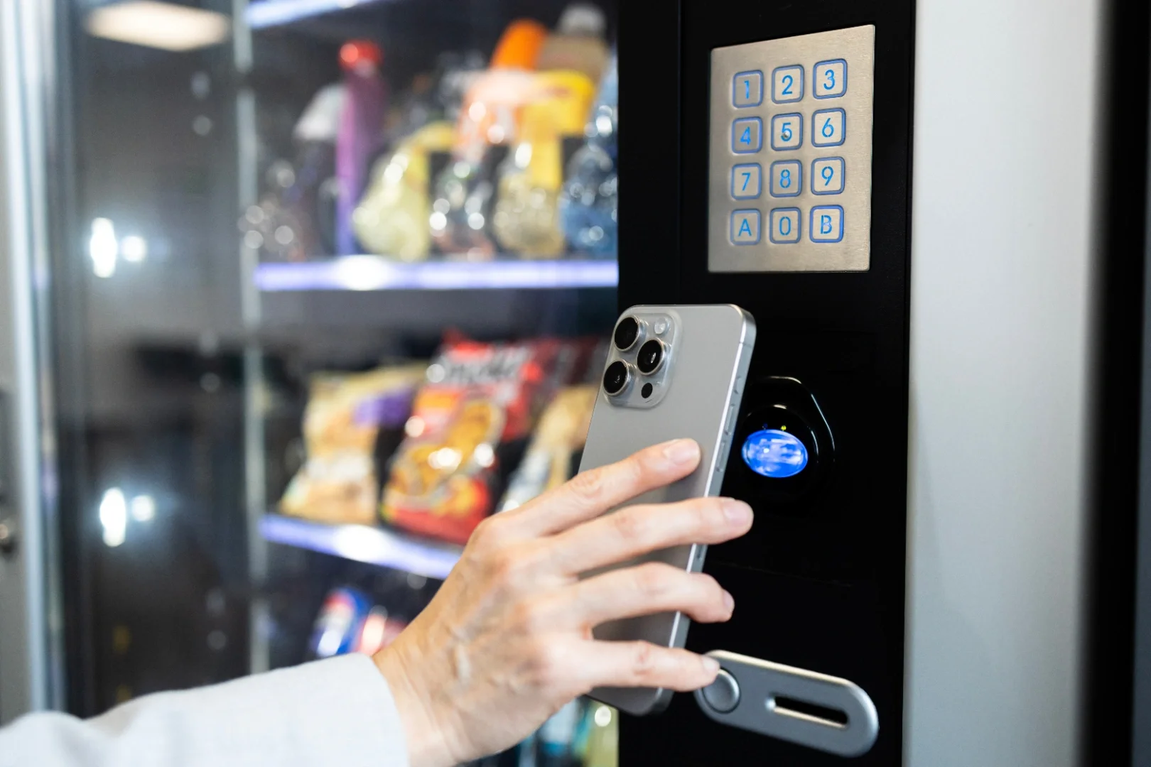 A hand holding a smartphone near a vending machine's card reader, with snacks visible inside the vending machine in the background.