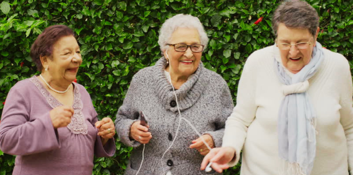 Three elderly women, smiling and laughing, standing outdoors against a green leafy background.