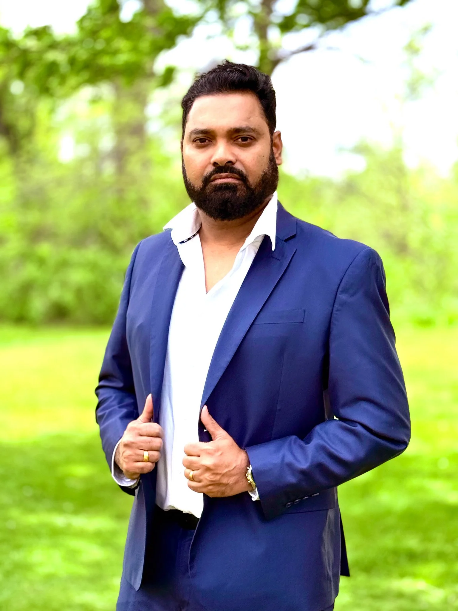 A man in a blue suit with a white shirt, with a beard and short dark hair, standing outdoors in a green natural setting, looking at the camera with a serious expression.