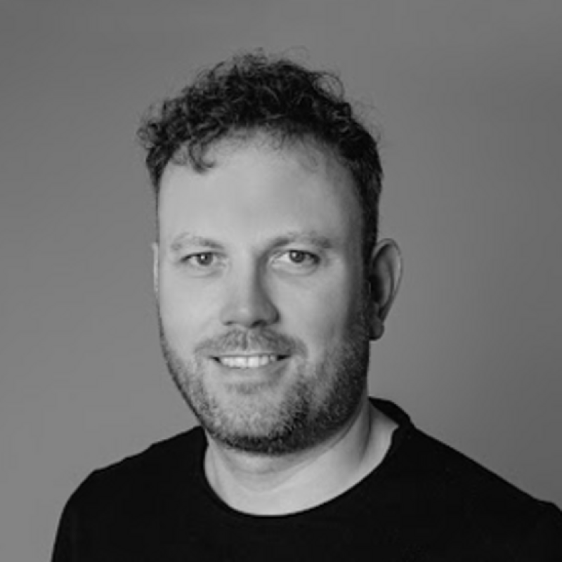 Black and white portrait of a man with short curly hair, a beard, and a mustache, wearing a dark t-shirt, smiling at the camera.