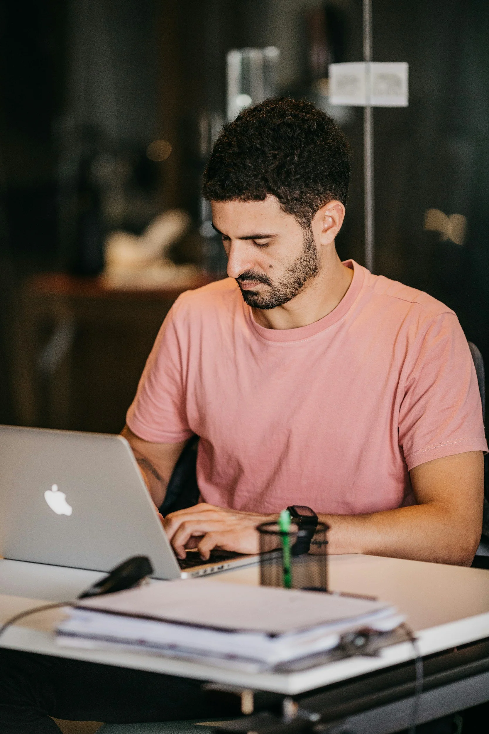 A man with dark hair and a short beard, wearing a salmon pink t-shirt, working on his laptop.