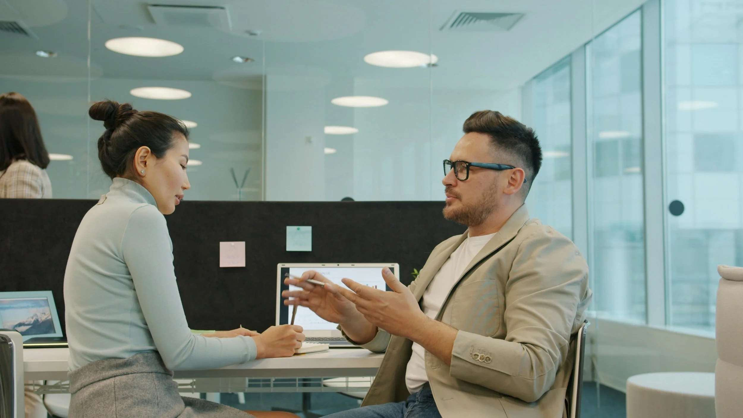 A white man wearing a beige blazer and glasses, talking to a female colleague in an office environment