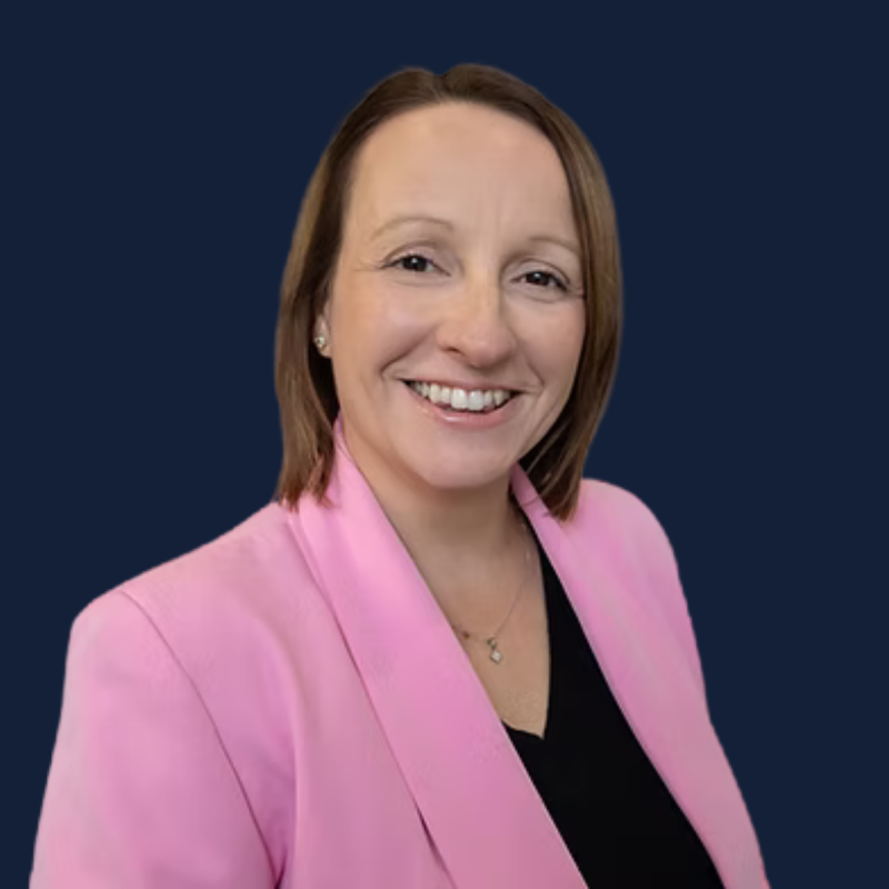 Portrait of a woman with shoulder-length brown hair, wearing a pink blazer over a black top, smiling against a dark blue background.