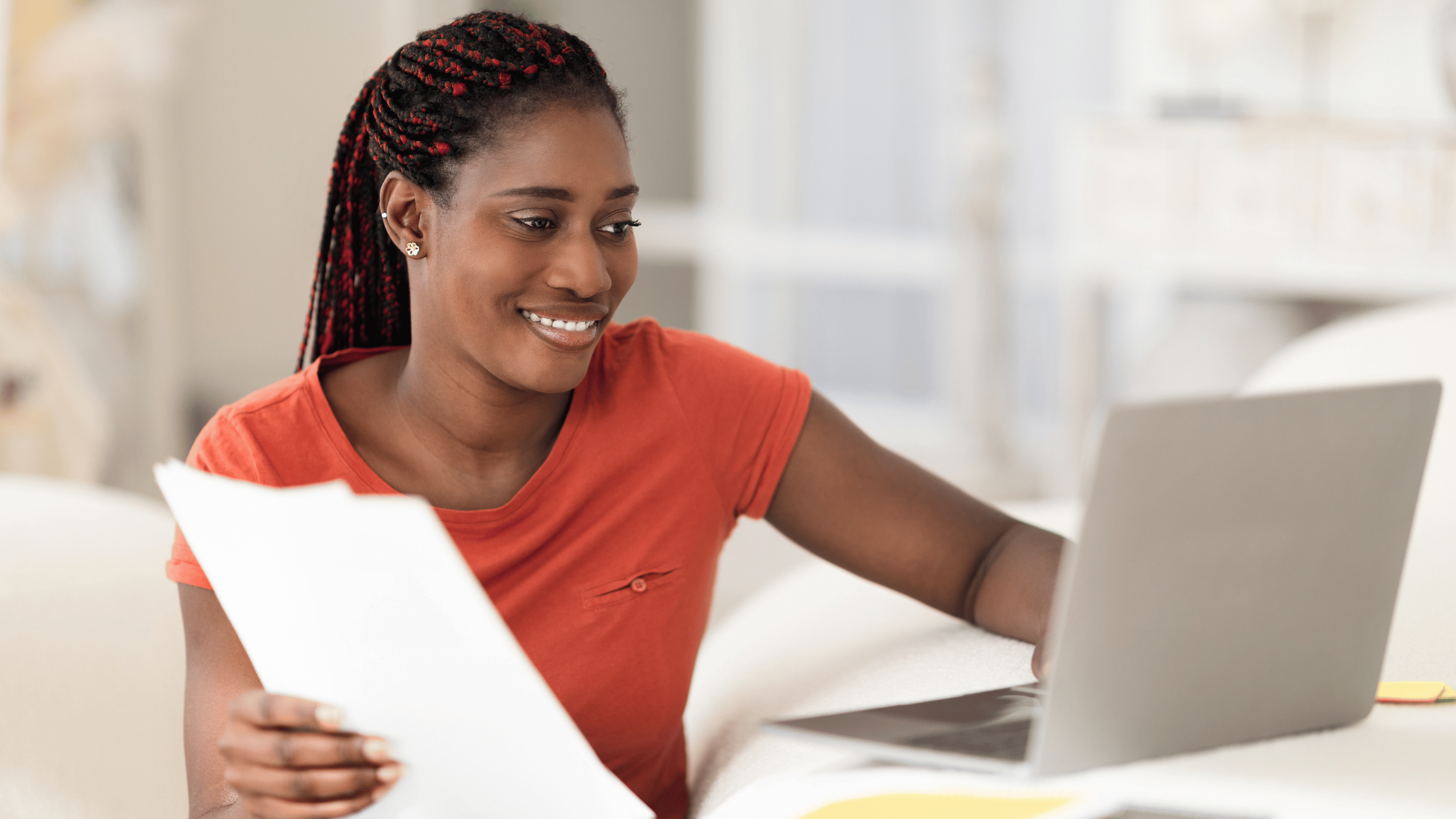 A black woman with long braided hair worn in a ponytail, wearing an organge t-shirt, holding some papers and working at her laptop.
