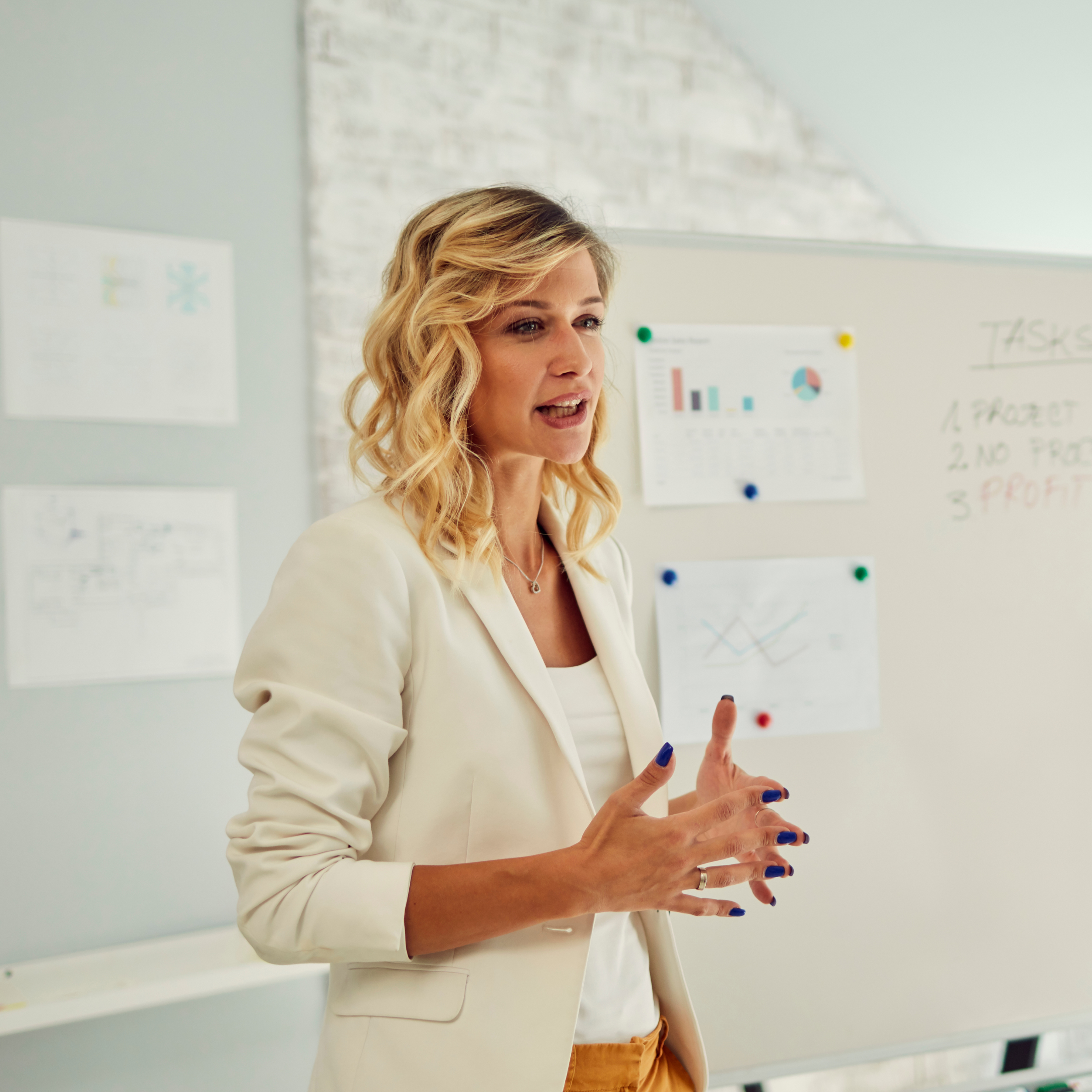 A woman with wavy blonde hair and a white blazer presenting in front of a whiteboard with graphs and notes in a room with a white brick wall.