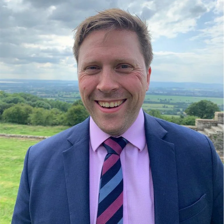 A smiling man in a suit and tie standing outdoors with a scenic landscape of greenery and sky in the background.