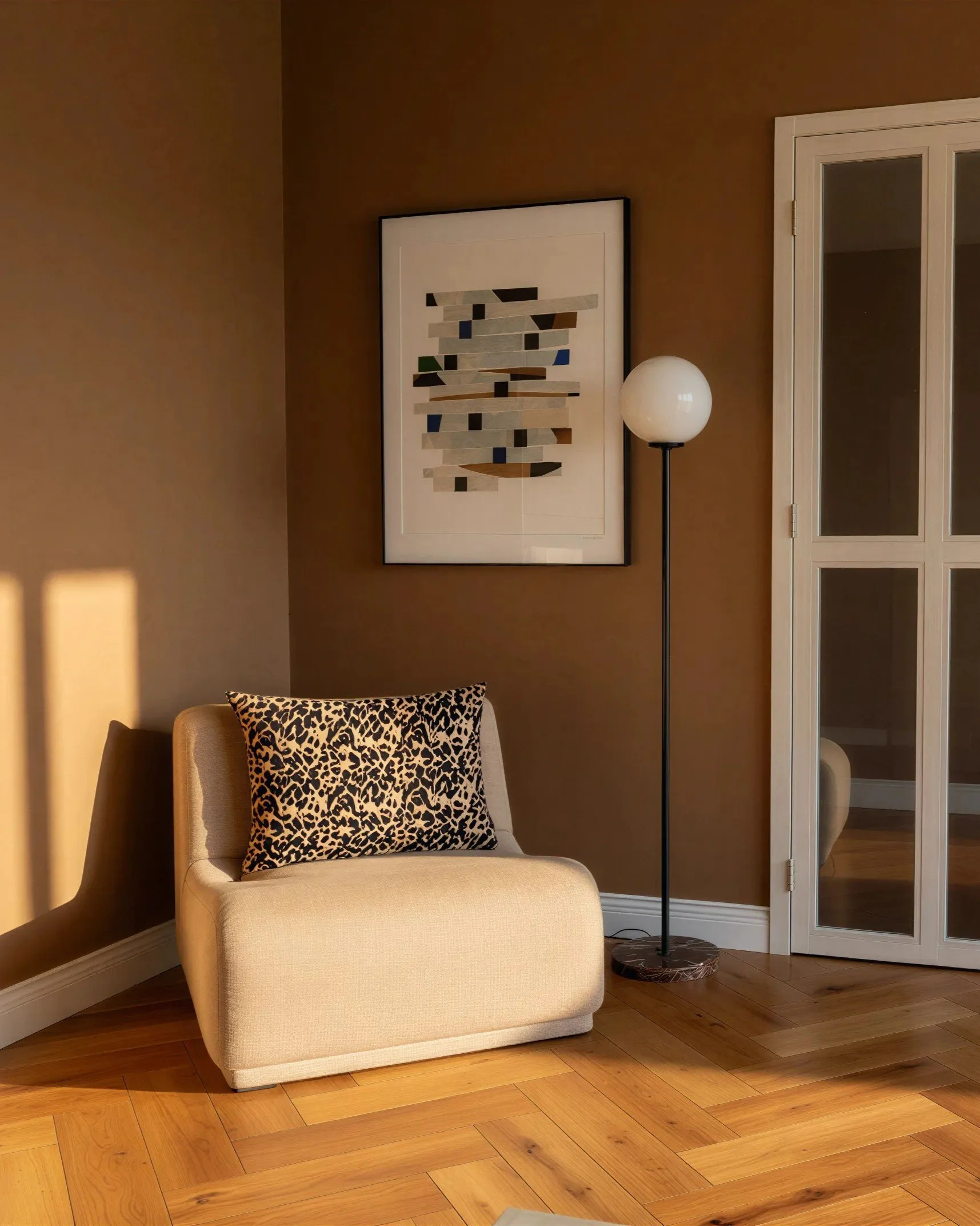 A beige armchair with a leopard print pillow, a tall black floor lamp with a white spherical shade, a framed abstract artwork on a brown wall, and a white cabinet with glass doors on a hardwood floor.