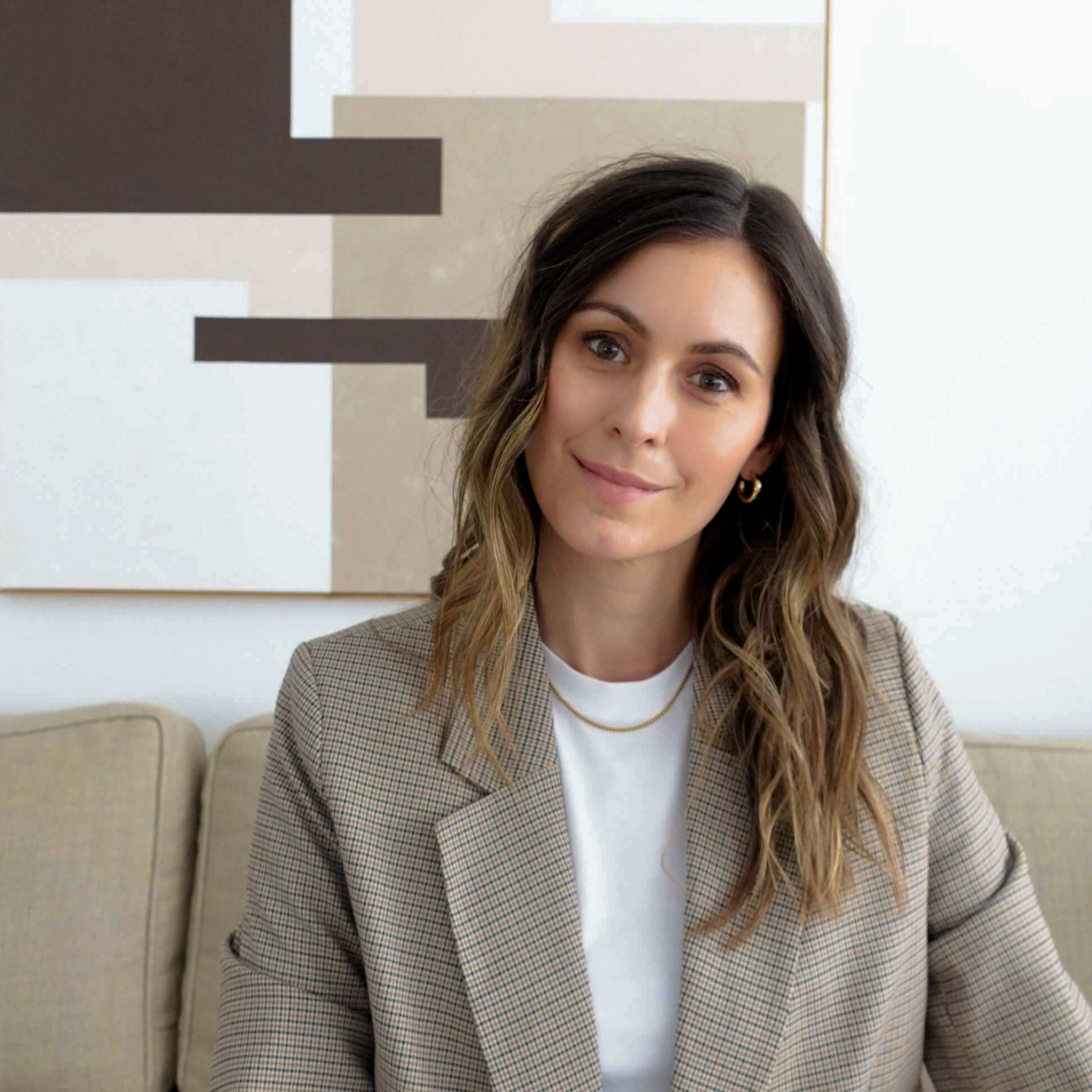 Close-up of a woman with long brown hair wearing hoop earrings, a white top, and a beige blazer with a checkered pattern, sitting in a room with beige chairs and abstract art on the wall.