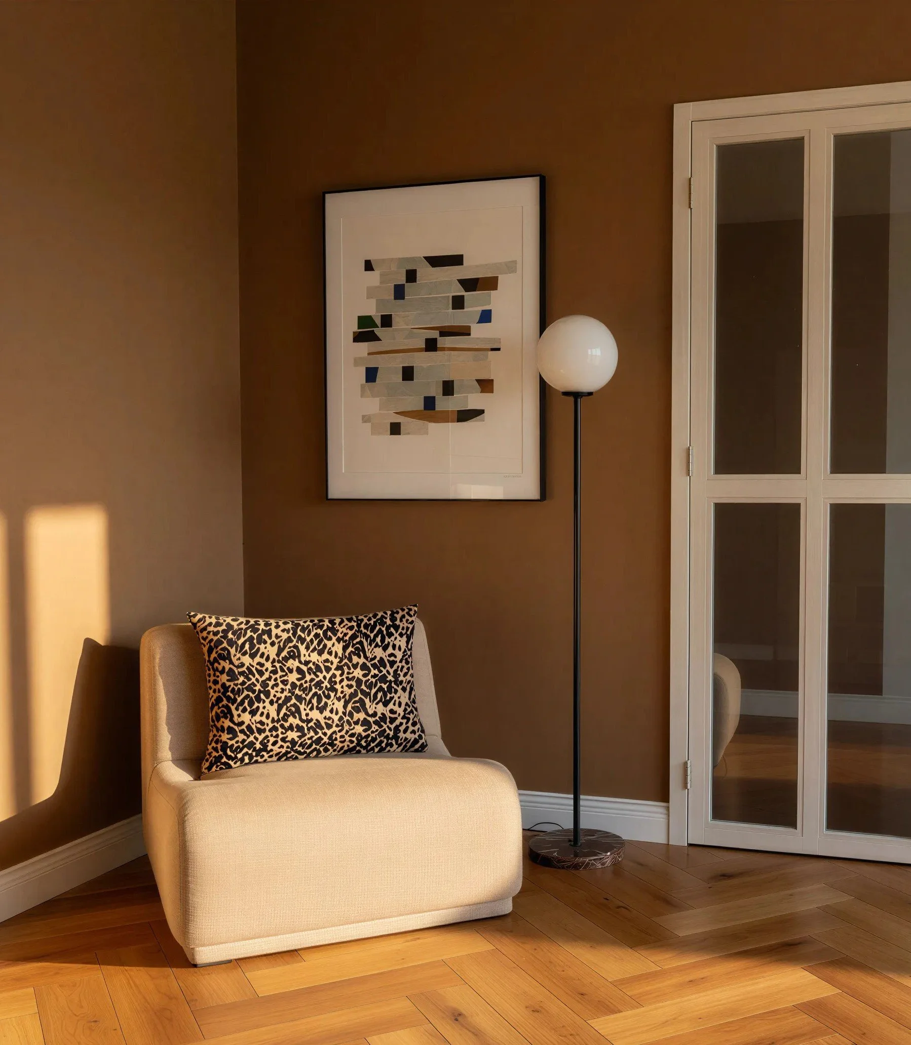 A beige armchair with a black and white leopard print pillow on it, positioned in front of a brown wall with a framed abstract artwork hanging above. To the right, a white cabinet with glass doors stands next to a tall black floor lamp with a white spherical lampshade. The floor is hardwood with a natural finish.