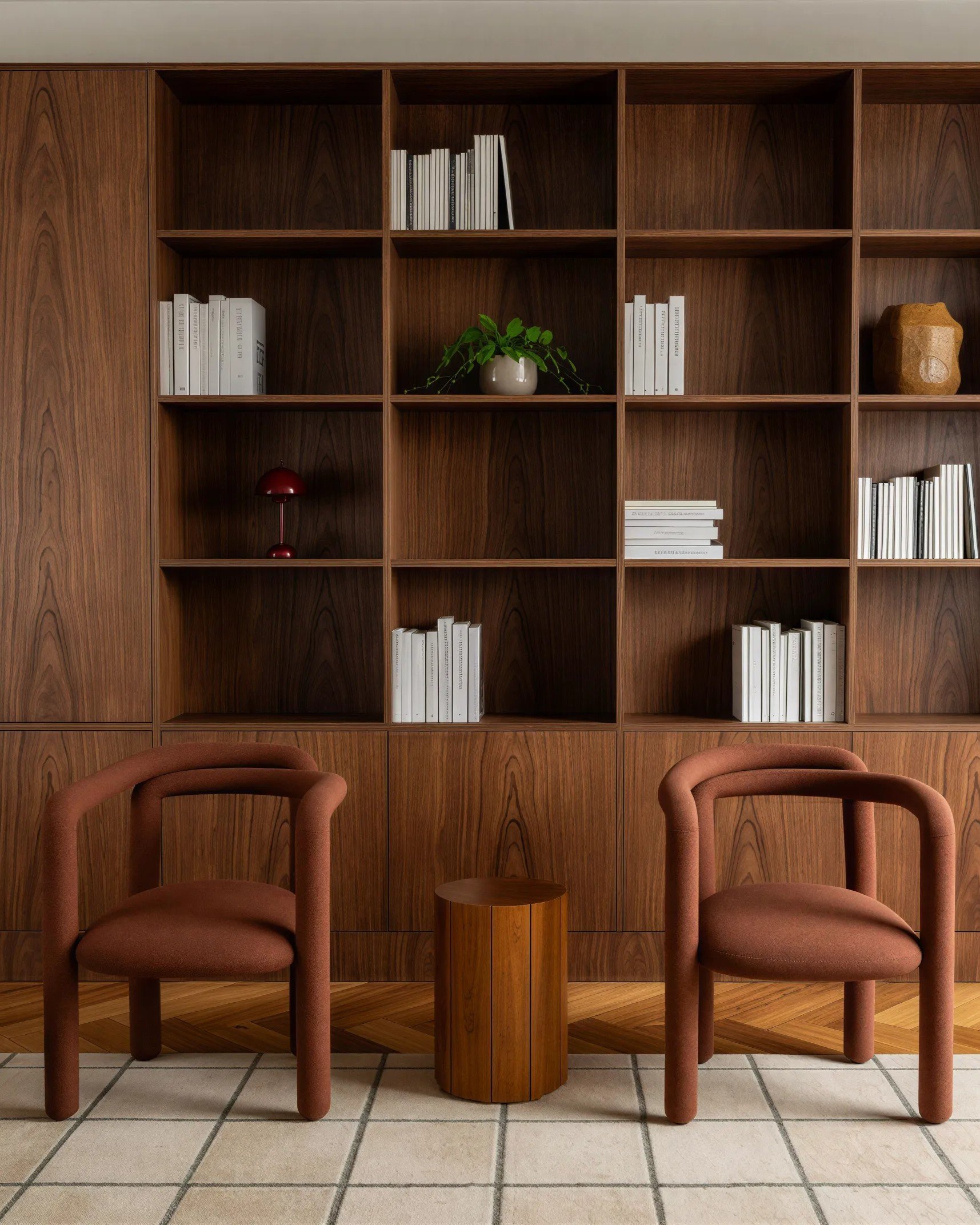 A wooden shelving unit with square compartments, some holding white books or magazines, a green potted plant in a white pot, a red table lamp, and a brown decorative object. In front are two pink upholstered chairs with rounded backs and curved armre