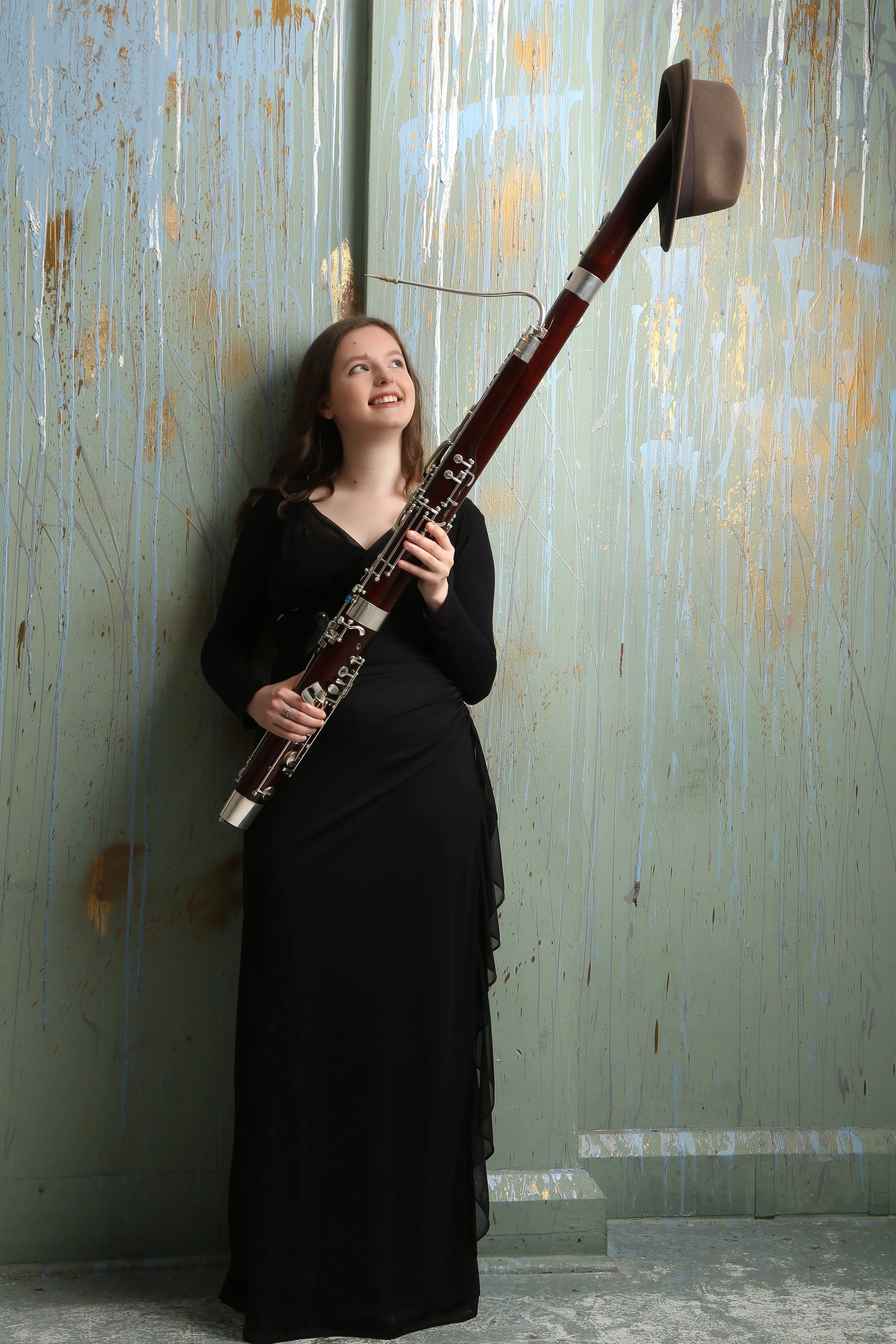 Young woman in a black dress leaning against a textured, splatter-painted wall, holding a bassoon and looking up smiling.