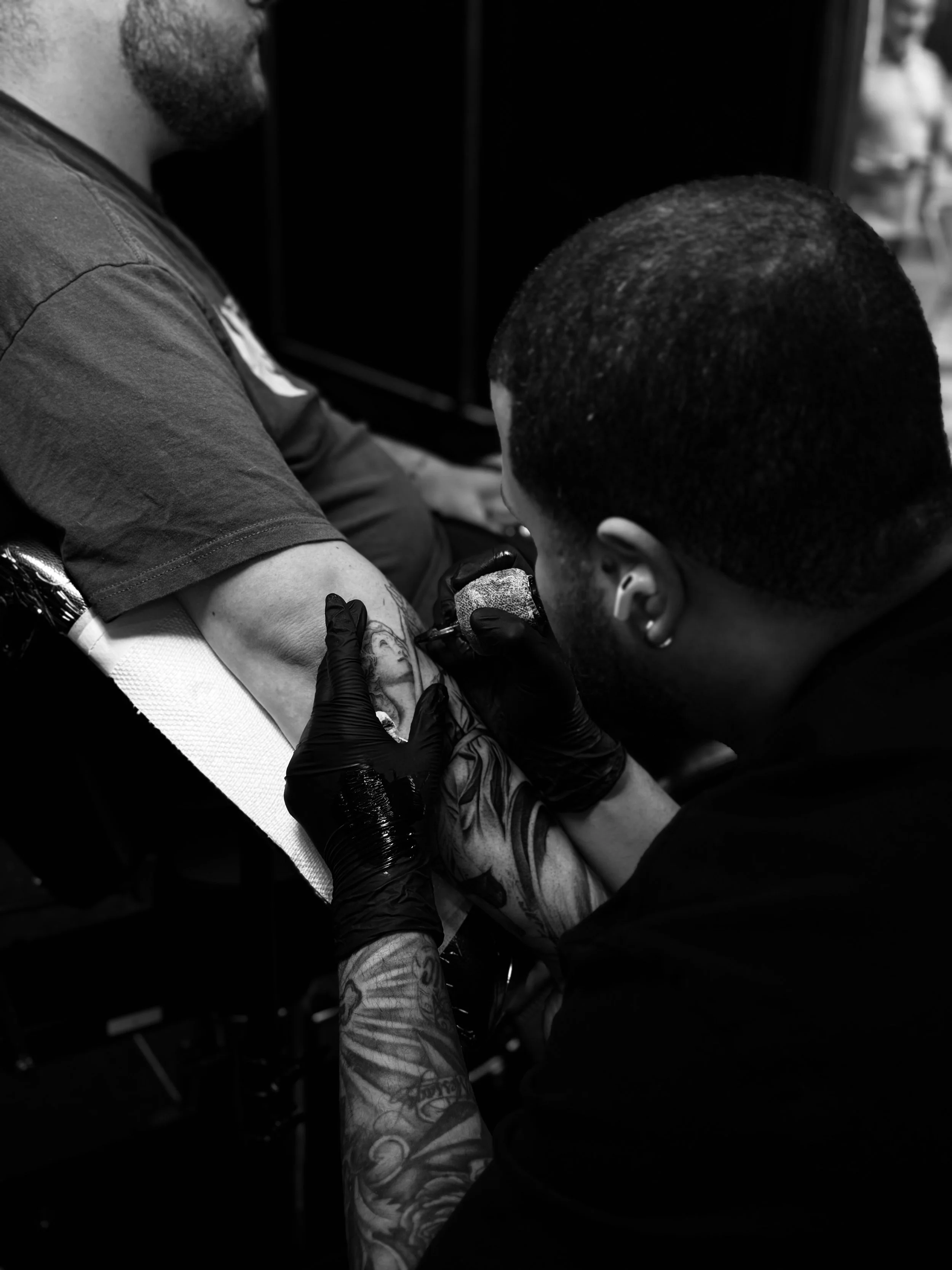 A man with tattoos on his arm receiving a tattoo on his forearm from a tattoo artist in black gloves, in a tattoo studio.