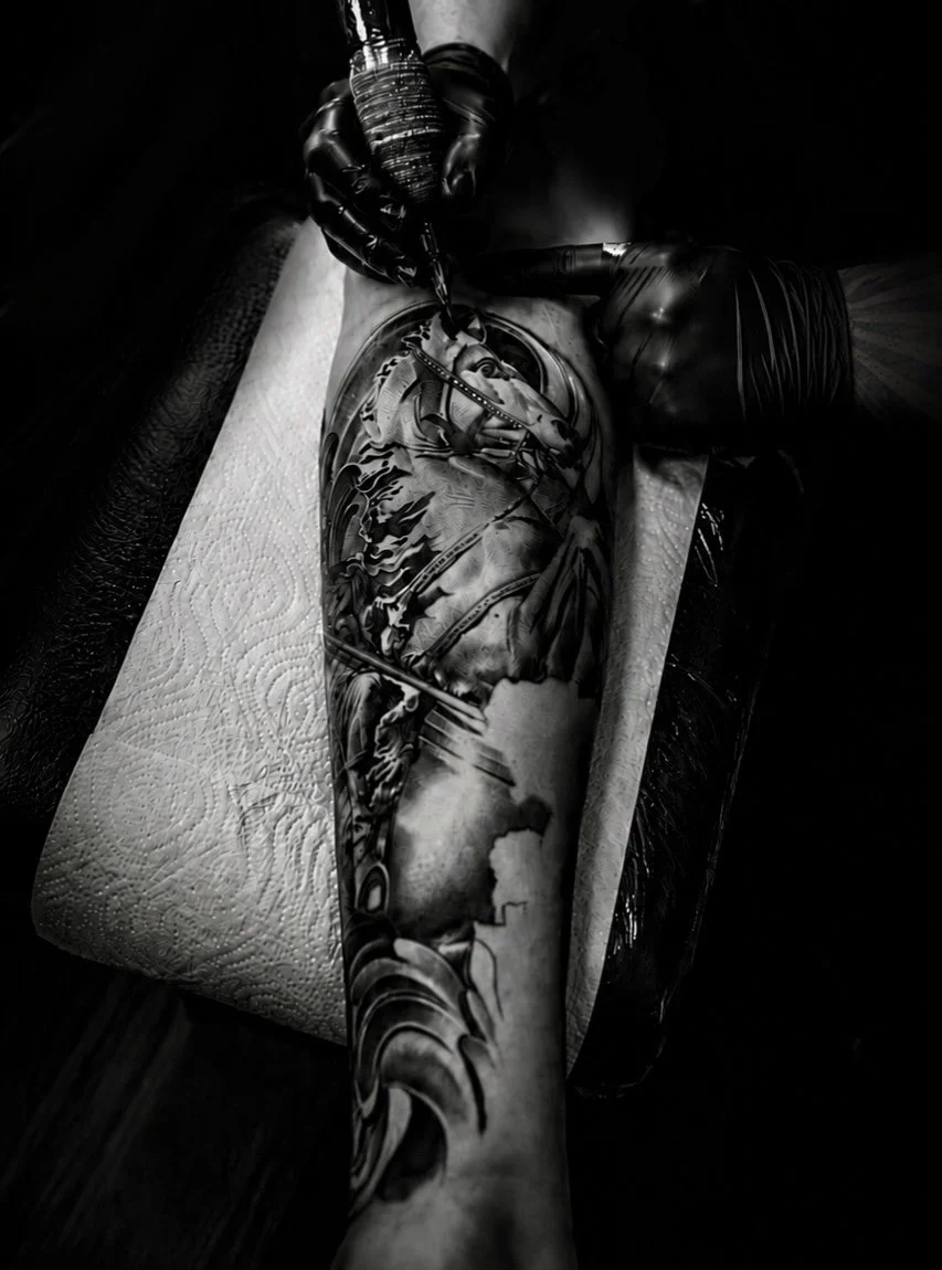 A person with black gloves is ink tattooing a detailed grayscale tattoo on another person's arm and forearm, depicting a horse and abstract patterns.