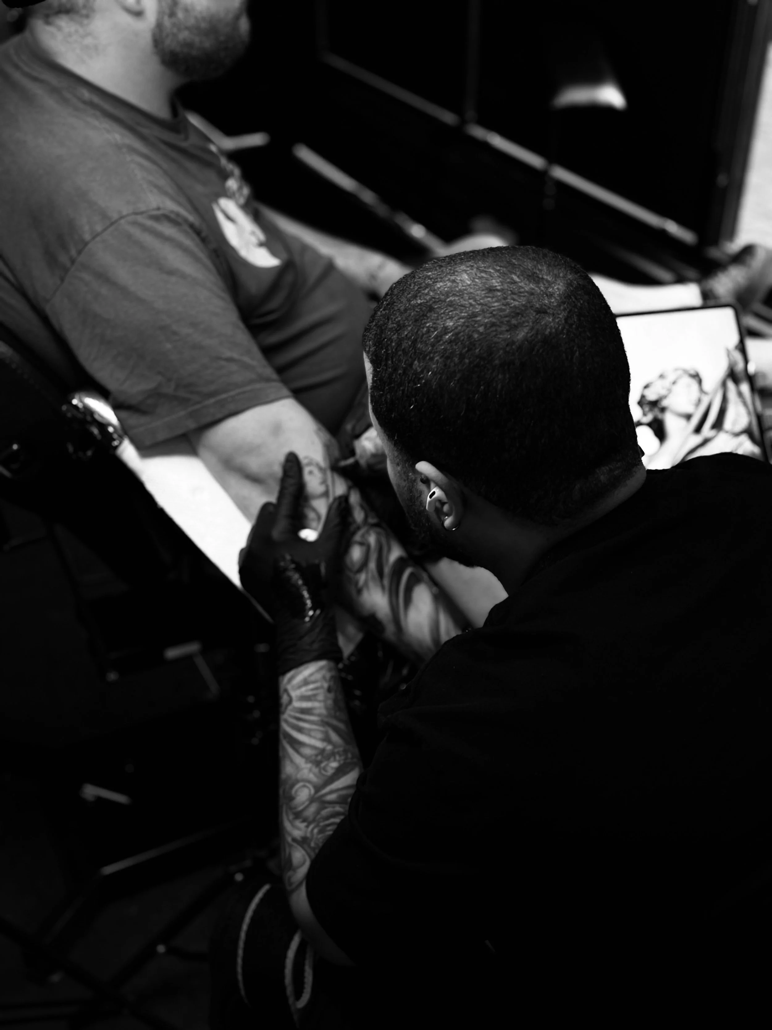 A person with short hair and tattoos on their arms is tattooing another person's arm. The person receiving the tattoo is sitting on a chair, wearing a t-shirt.