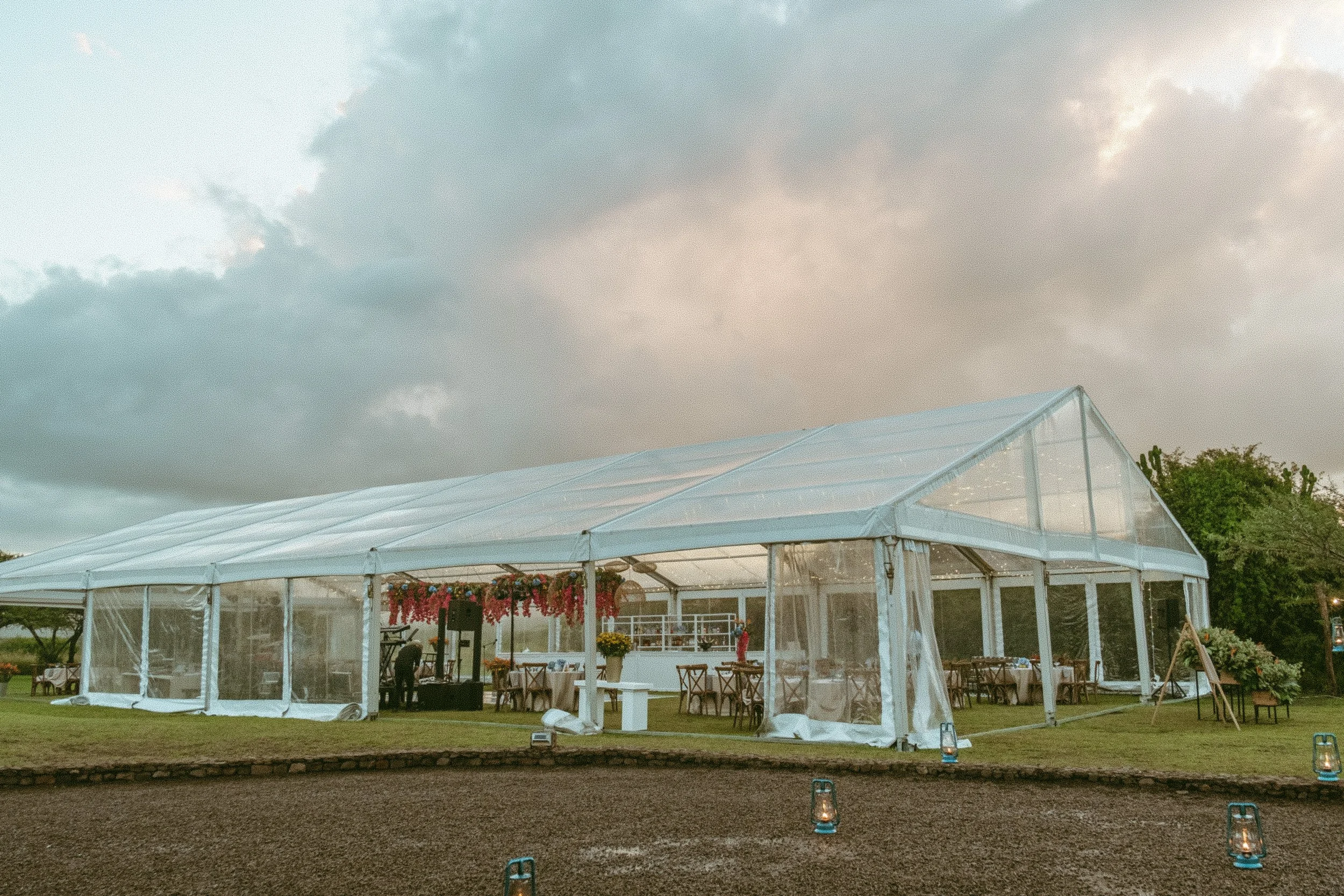 A large white event tent with clear side panels set up outdoors, decorated with pink flowers and surrounded by tables and chairs, with a cloudy sky overhead and small lanterns on the ground.
