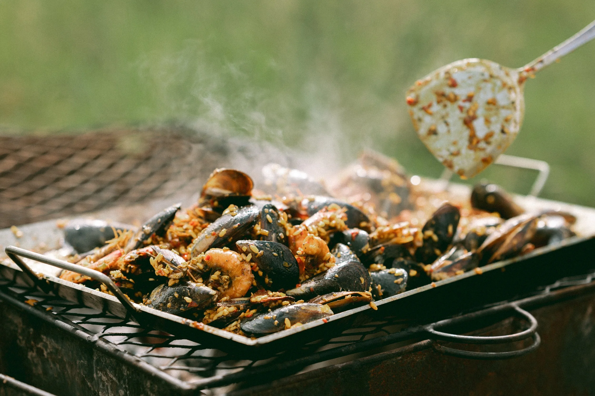 Cooked seafood, including mussels and shrimp, being grilled over an open flame with rice and spices, on a black grill.