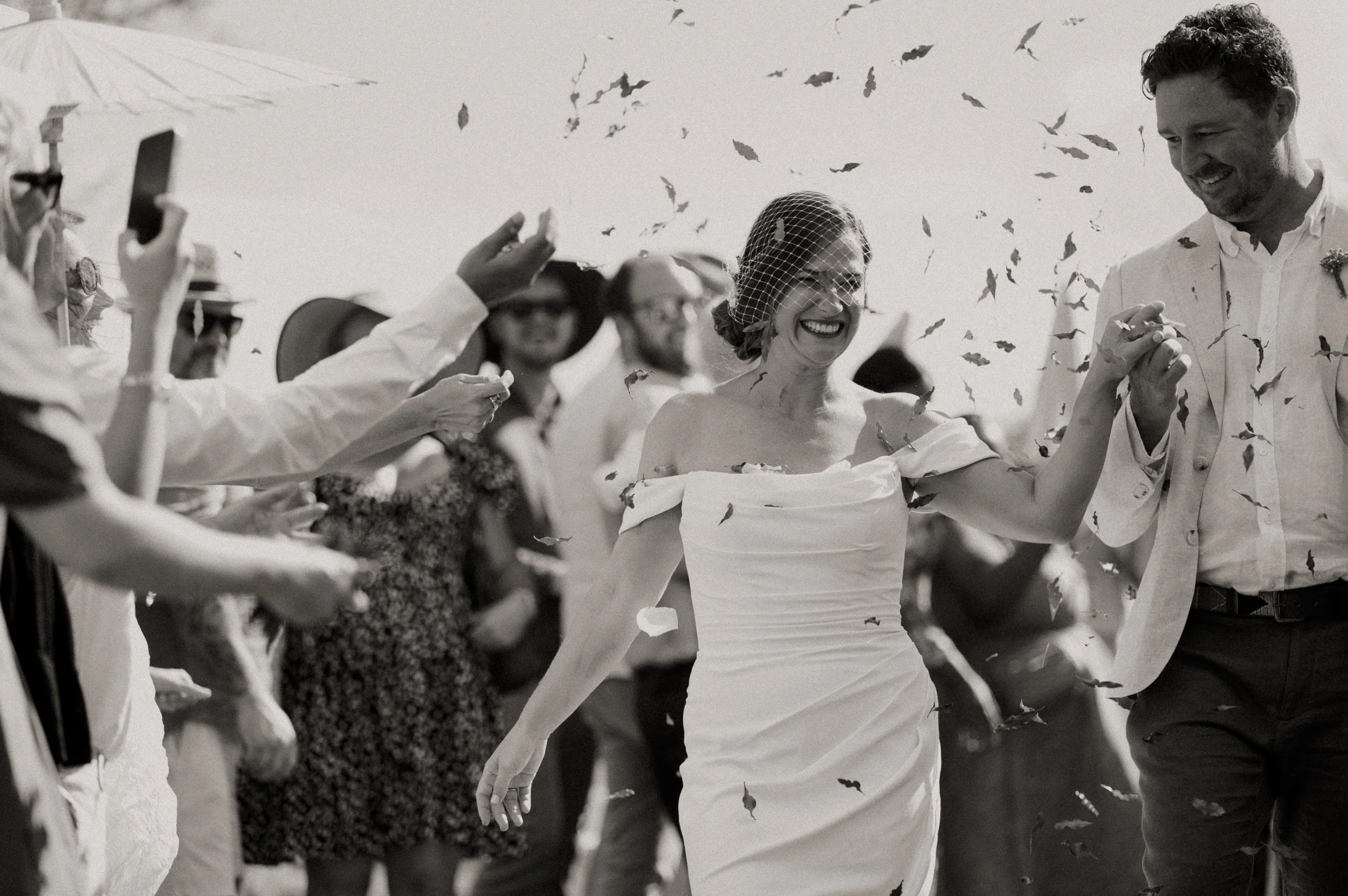 A bride and groom hold hands and smile at each other during their wedding celebration, surrounded by guests and falling confetti.
