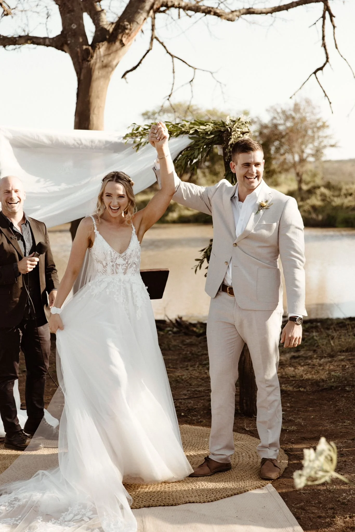 A wedding ceremony outdoors with a bride and groom holding hands and smiling, surrounded by trees and natural scenery.