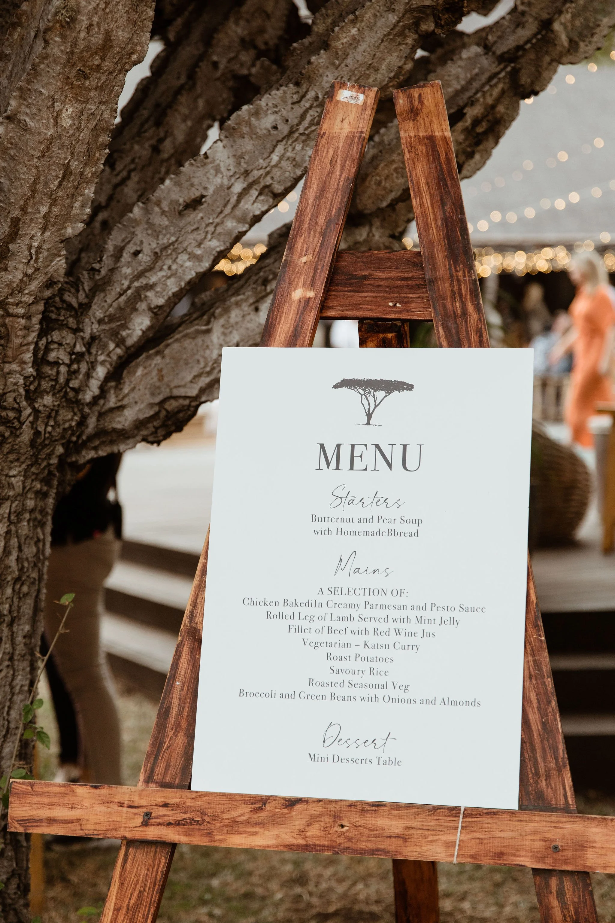A menu sign displayed on a wooden easel outdoors, next to a tree with a bark texture. The menu features starters, mains, and desserts, and is part of an event or restaurant setting with blurry lights and people in the background.