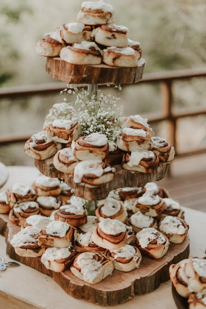 A multi-tiered wooden cupcake stand filled with cinnamon rolls topped with icing, decorated with small white flowers, on a rustic table.