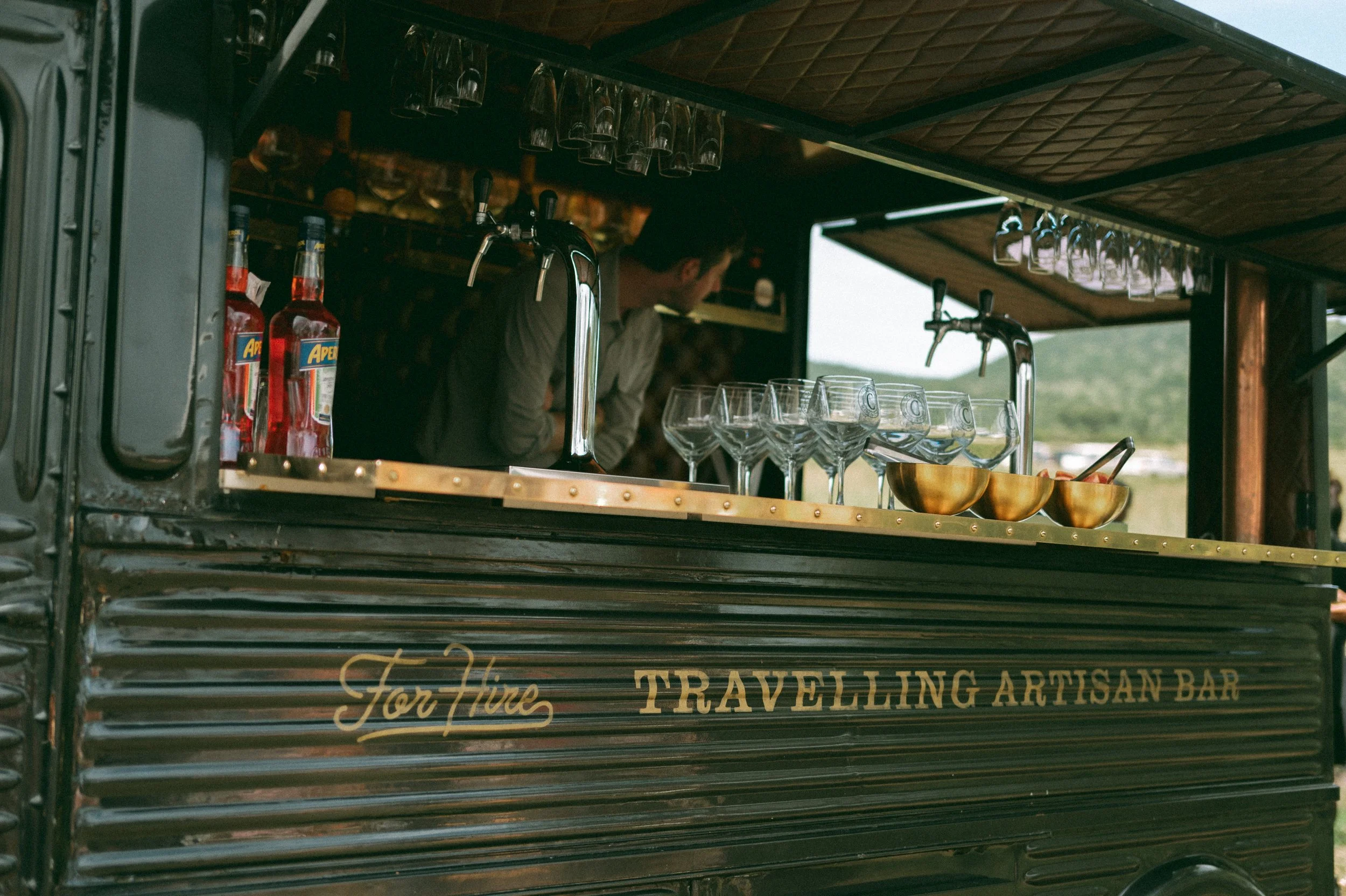 A travel artisan bar cart with glasses hanging upside down, bottles, and bowls, and a person behind the bar.