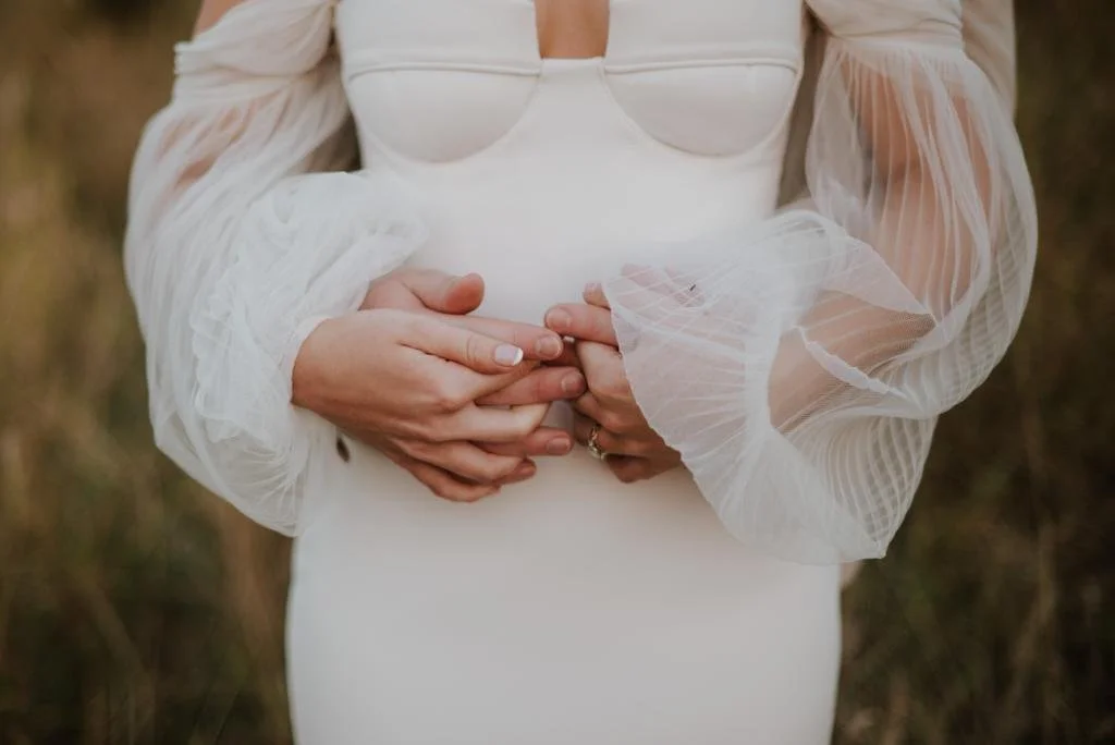 Woman wearing a white dress with sheer, puffed sleeves, holding her pregnant belly with both hands in an outdoor setting.