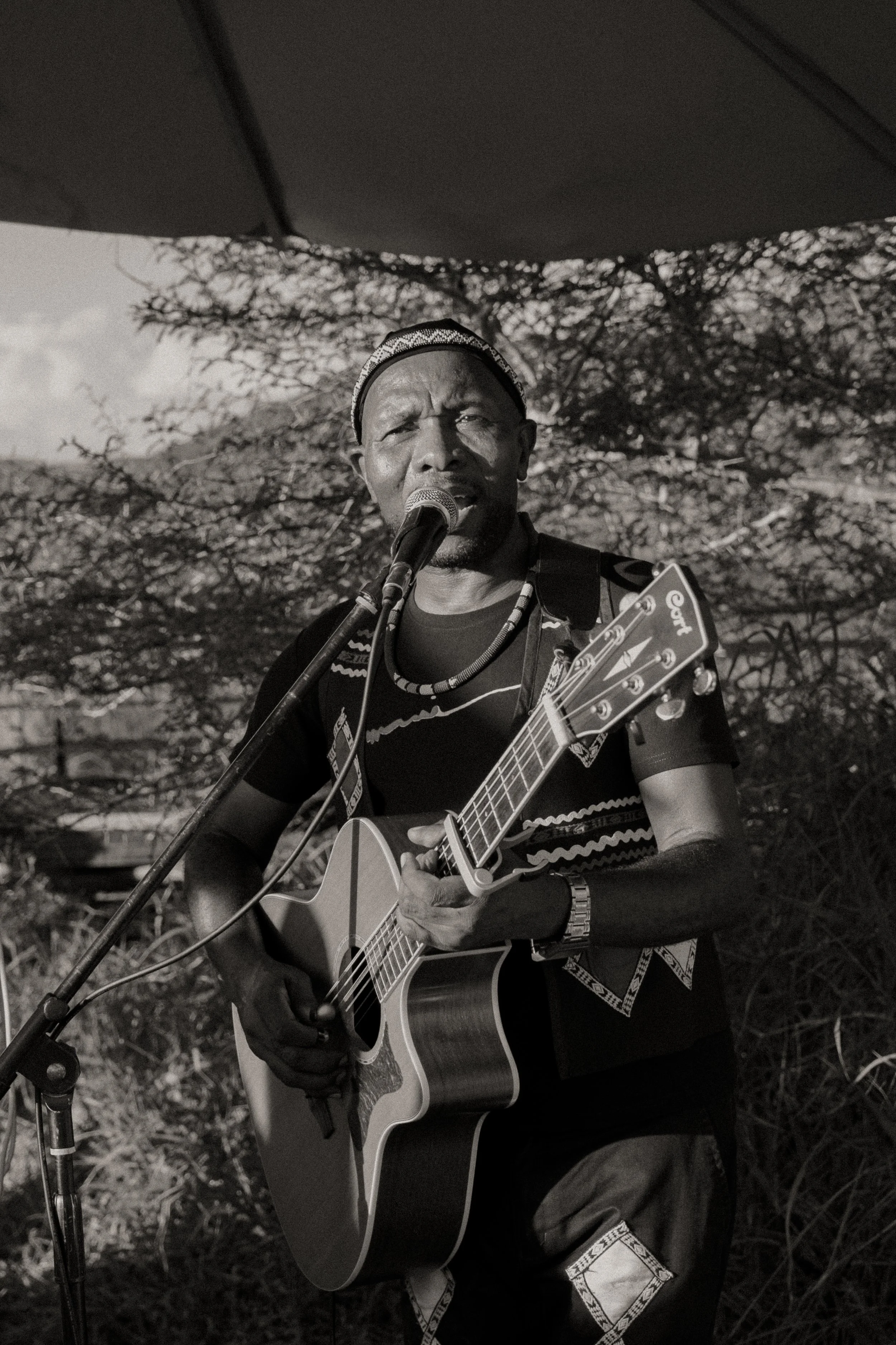 A man performing outdoors, playing an acoustic guitar and singing into a microphone, wearing a patterned shirt, a beaded necklace, a wristwatch, and a patterned headwrap, with trees and a cloudy sky in the background.