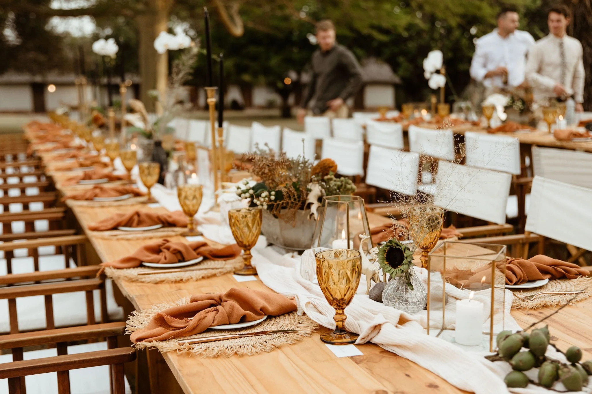 An outdoor dining table set for a celebration, decorated with flowers, candles, and tableware, with trees in the background.
