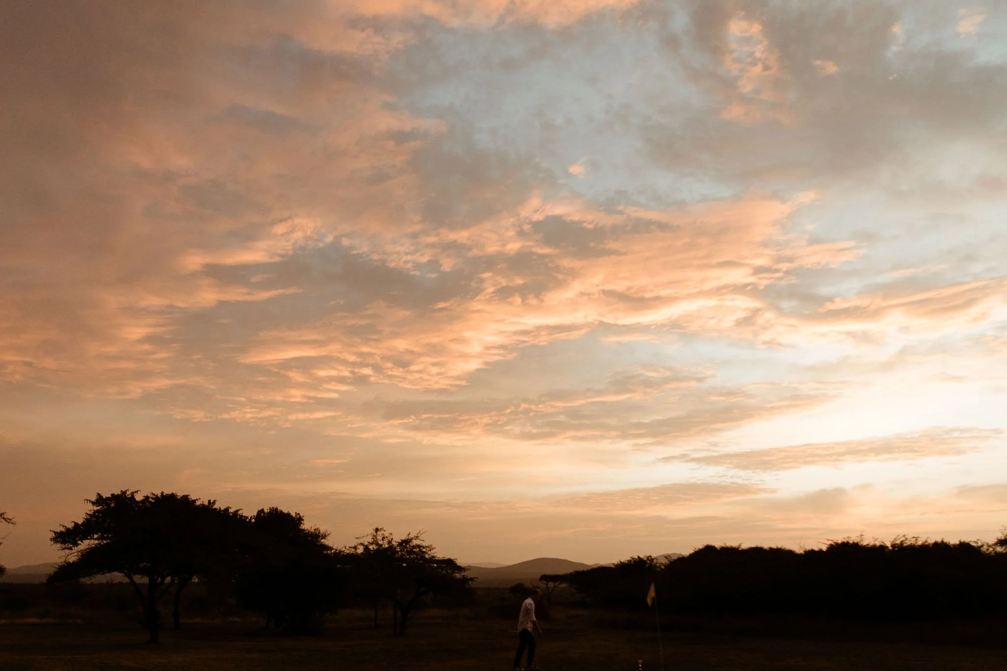 A landscape during sunset showing orange and pink clouds in the sky, silhouetted trees, and a person walking in the foreground.