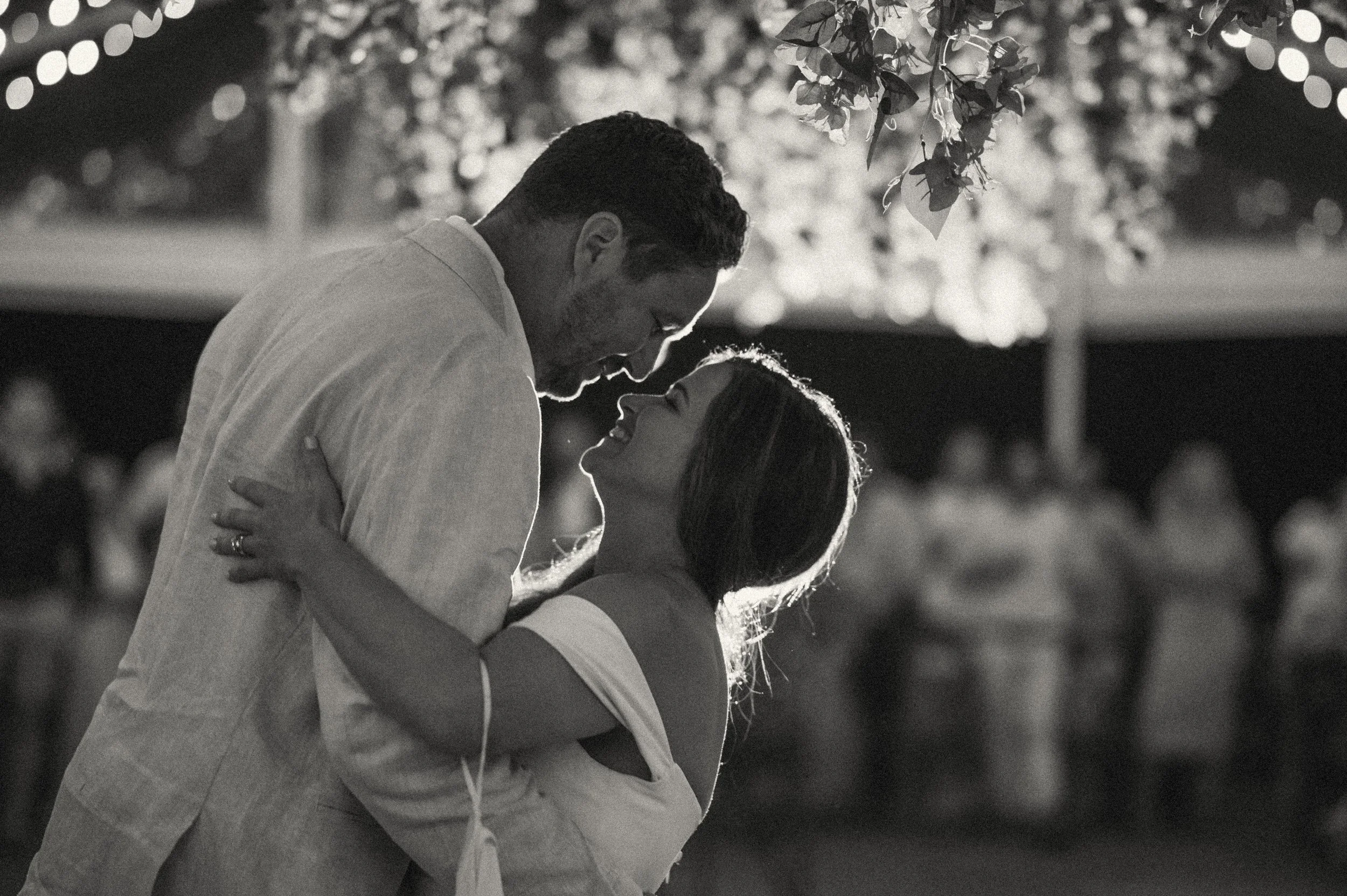 A black and white photo of a couple dancing intimately outdoors, with the man wearing a suit and the woman in a sleeveless dress, smiling and looking at each other, surrounded by blurred lights and trees.