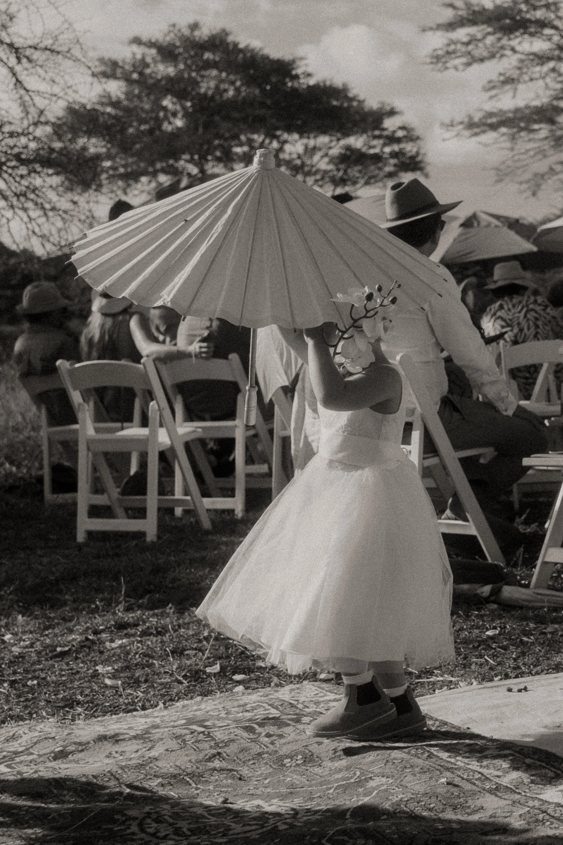 A flowergirl in a white dress and hat holding an umbrella and a branch with flowers, walking down the asile outdoors at a gathering with seated people, trees in the background, in black and white.