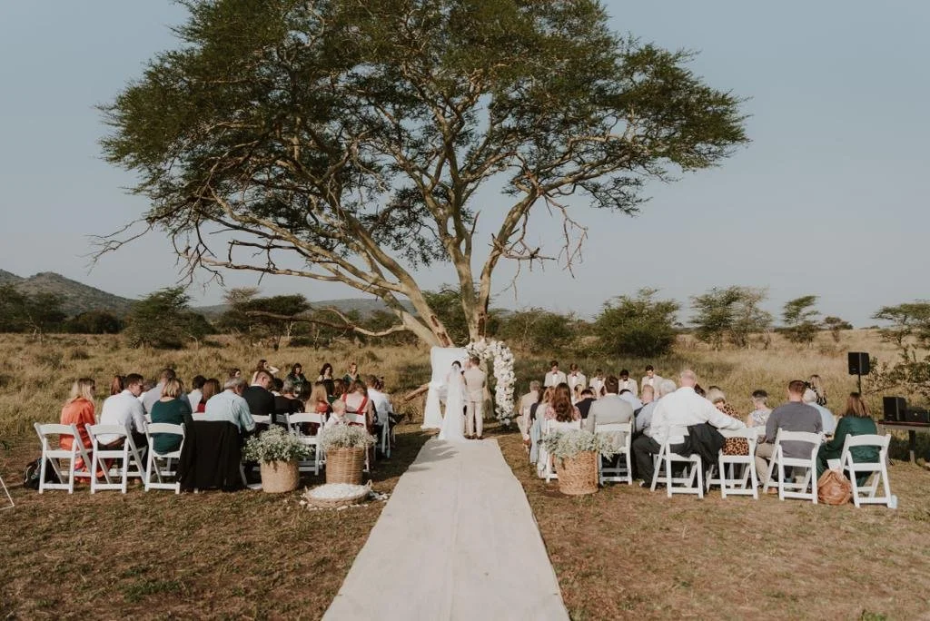 An outdoor wedding ceremony taking place beneath a large tree, with guests seated on both sides of a white aisle, and the bride and groom standing at the altar in a natural, open field setting.
