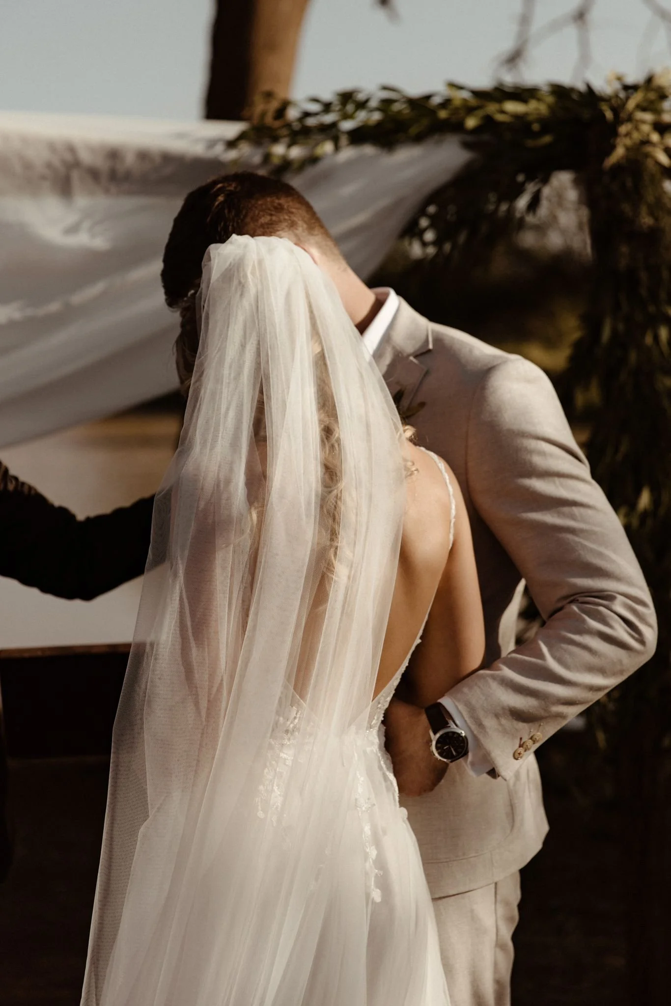 A bride and groom kissing, with the bride wearing a white wedding dress and veil, and the groom in a light-colored suit, in an indoor setting with soft lighting.