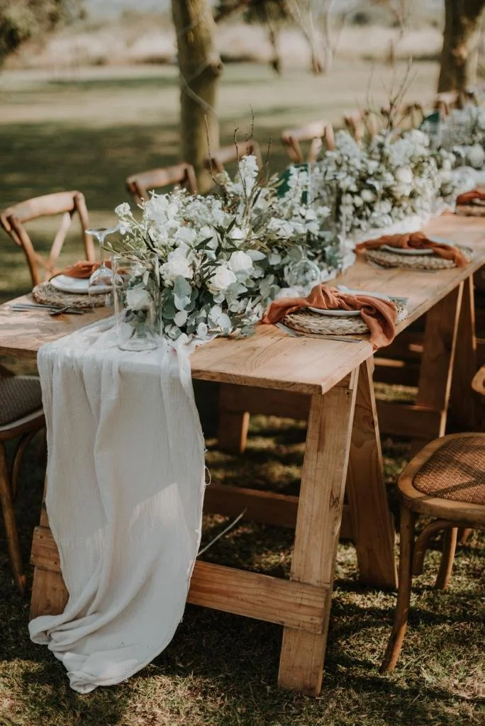Long wooden outdoor table decorated with white floral arrangements, greenery, and tableware, set by a river with trees in the background.