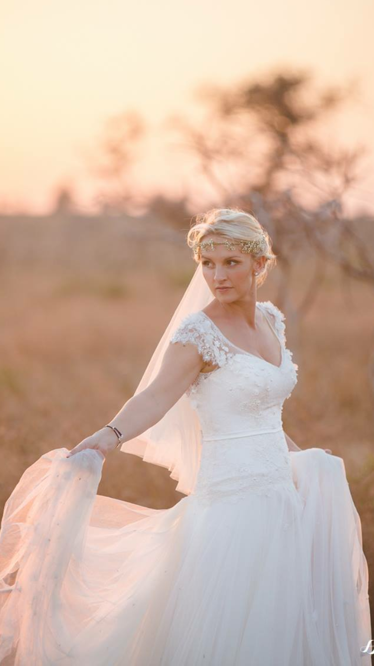 Bride in a white wedding dress with lace details, wearing a floral headband, standing outdoors at sunset with blurred trees in the background.