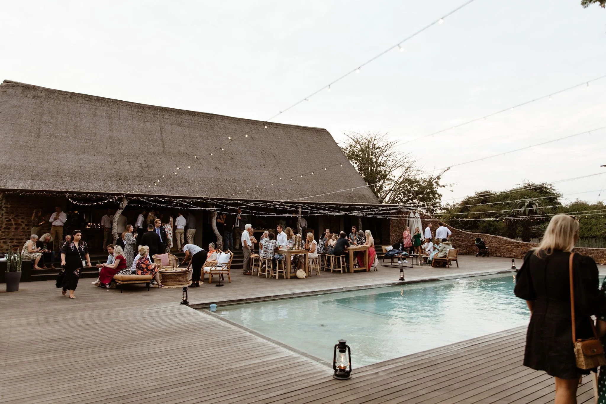 People gathered outdoors near a swimming pool, socializing and sitting at tables under string lights, with a building and trees in the background during early evening.