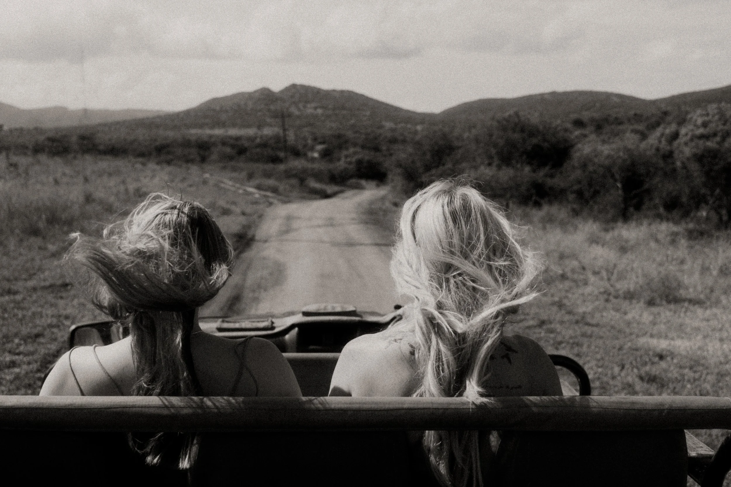 Two women with long hair riding in an open vehicle on a rural dirt road surrounded by hills and cloudy sky.