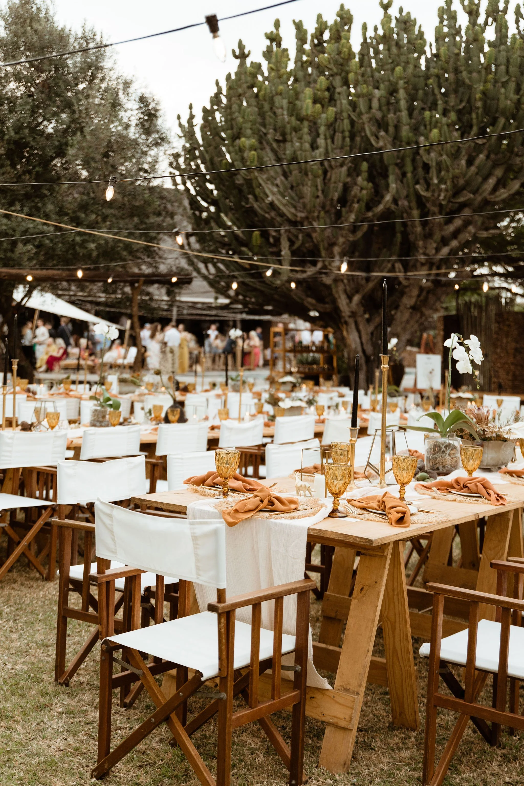 Outdoor event space with tables decorated with orange napkins, gold glasses, and floral centerpieces, set up under a large tree with string lights overhead and guests mingling in the background.