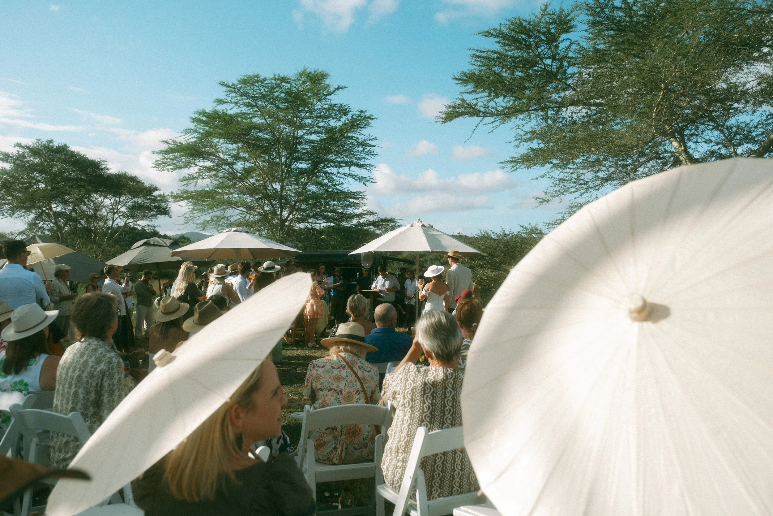 A large outdoor gathering with many people wearing summer clothing and hats, sitting and standing under umbrellas and trees during the daytime.